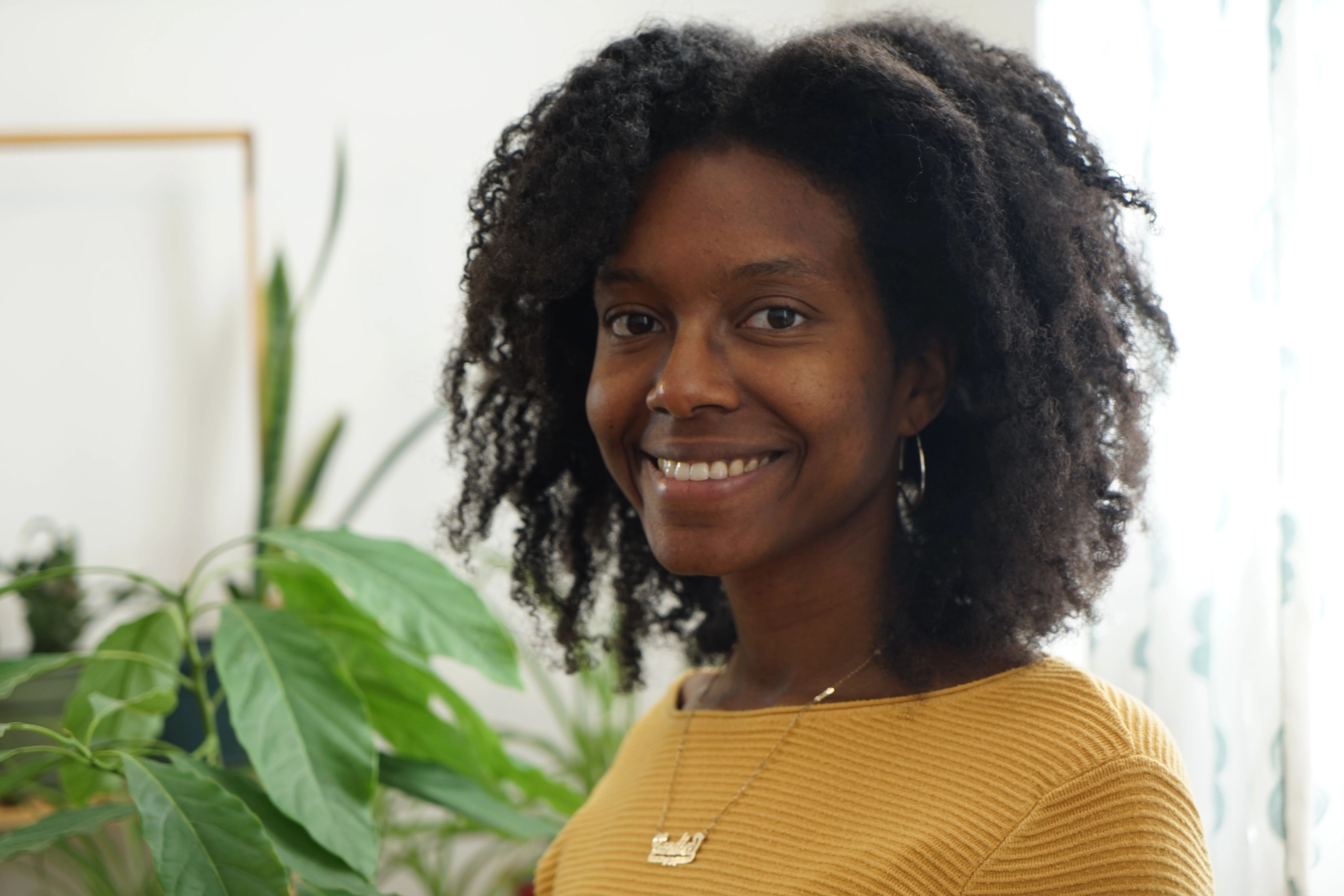 A smiling Black woman with curly hair looks at the camera, with plants in the background.