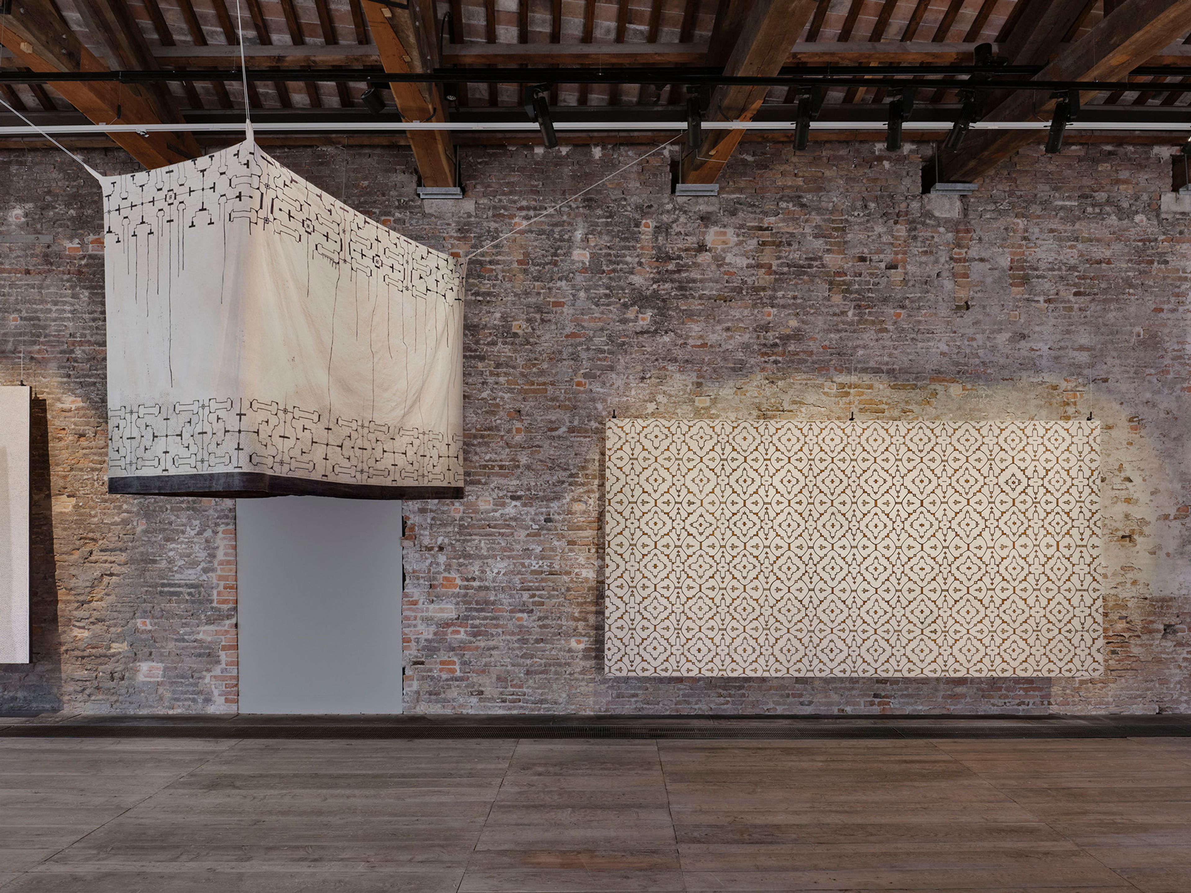 An art exhibit featuring a rectangular white fabric canopy with black patterns hanging from the ceiling, and a large rectangular patterned tapestry on a brick wall.
