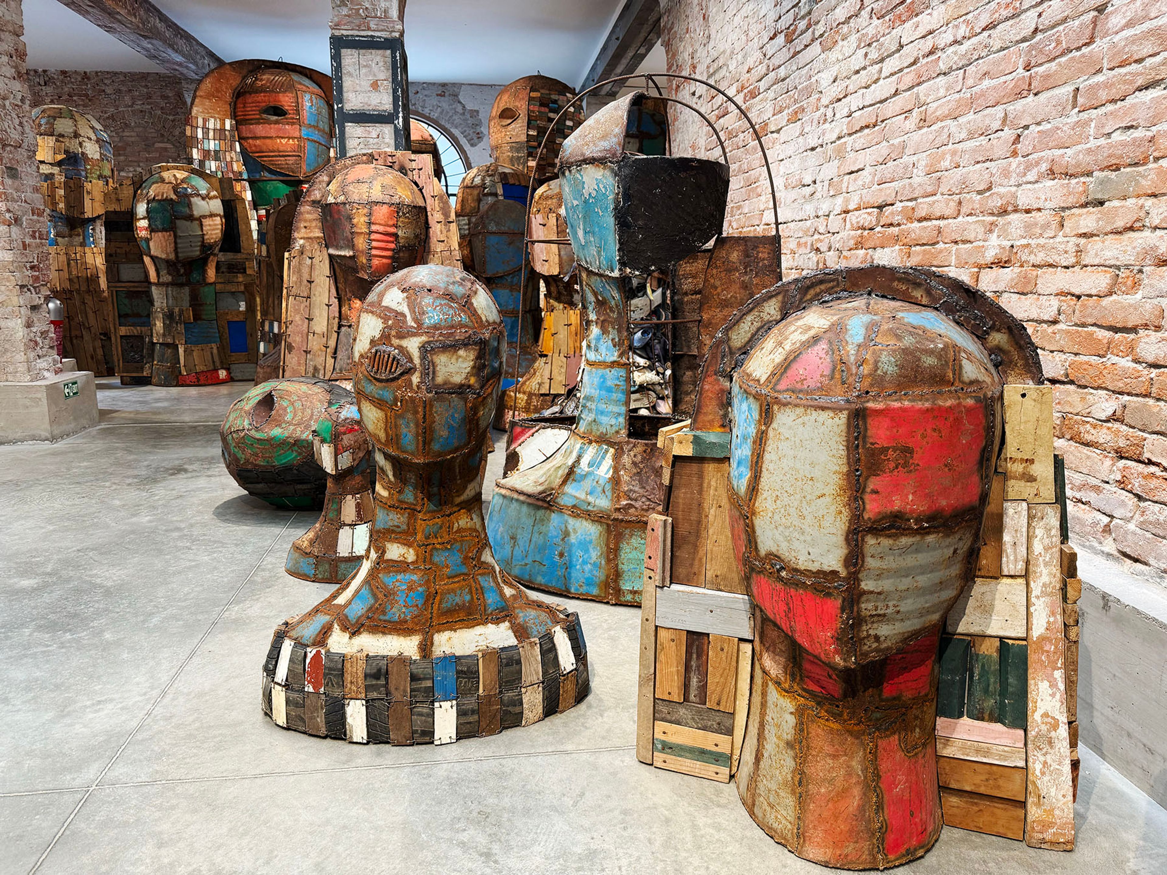 Numerous large, abstract human-like sculptures made from patchwork rusty metal and wood in an industrial gallery.