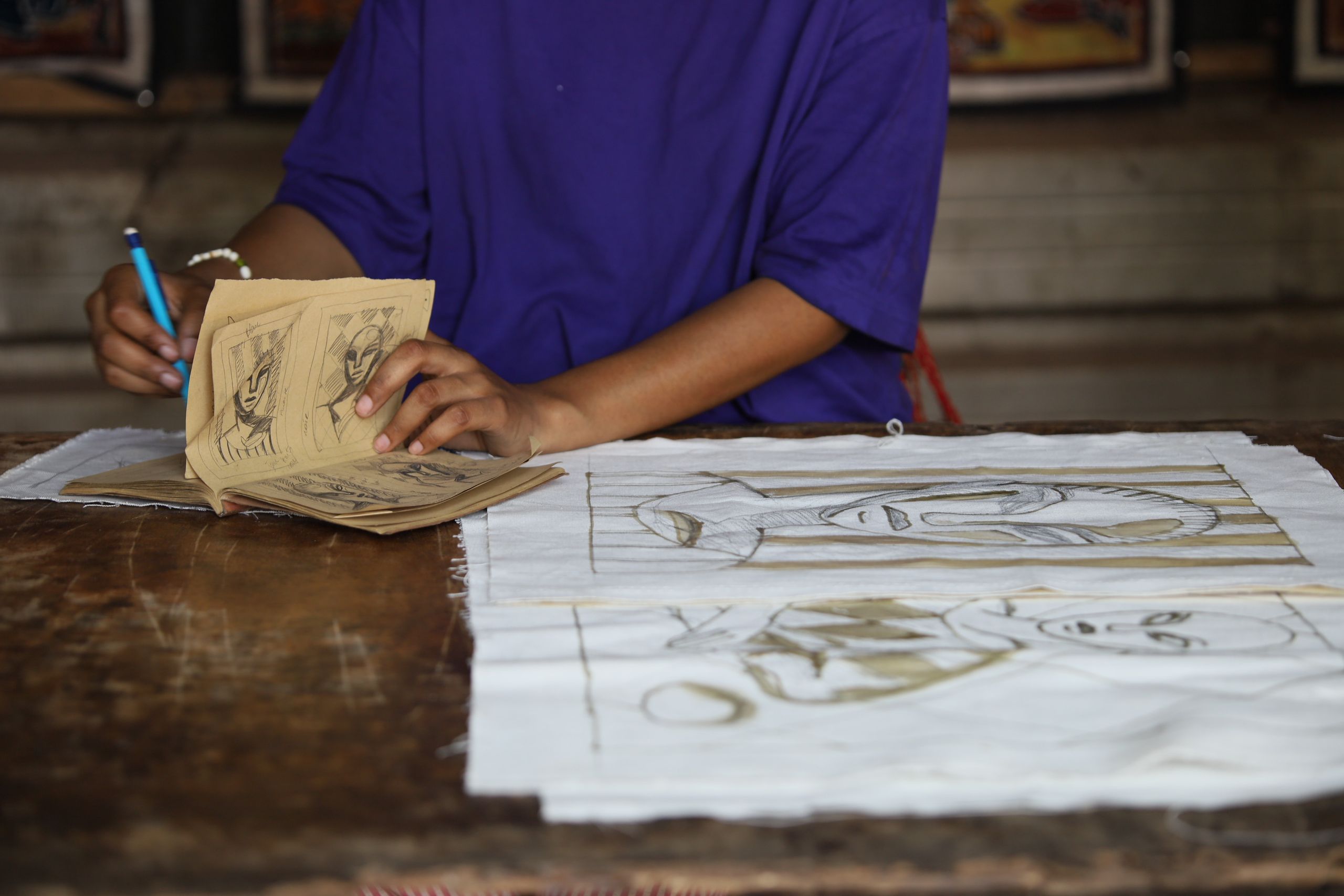 A person in a purple shirt looks at a sketchbook while working on abstract face drawings on fabric.