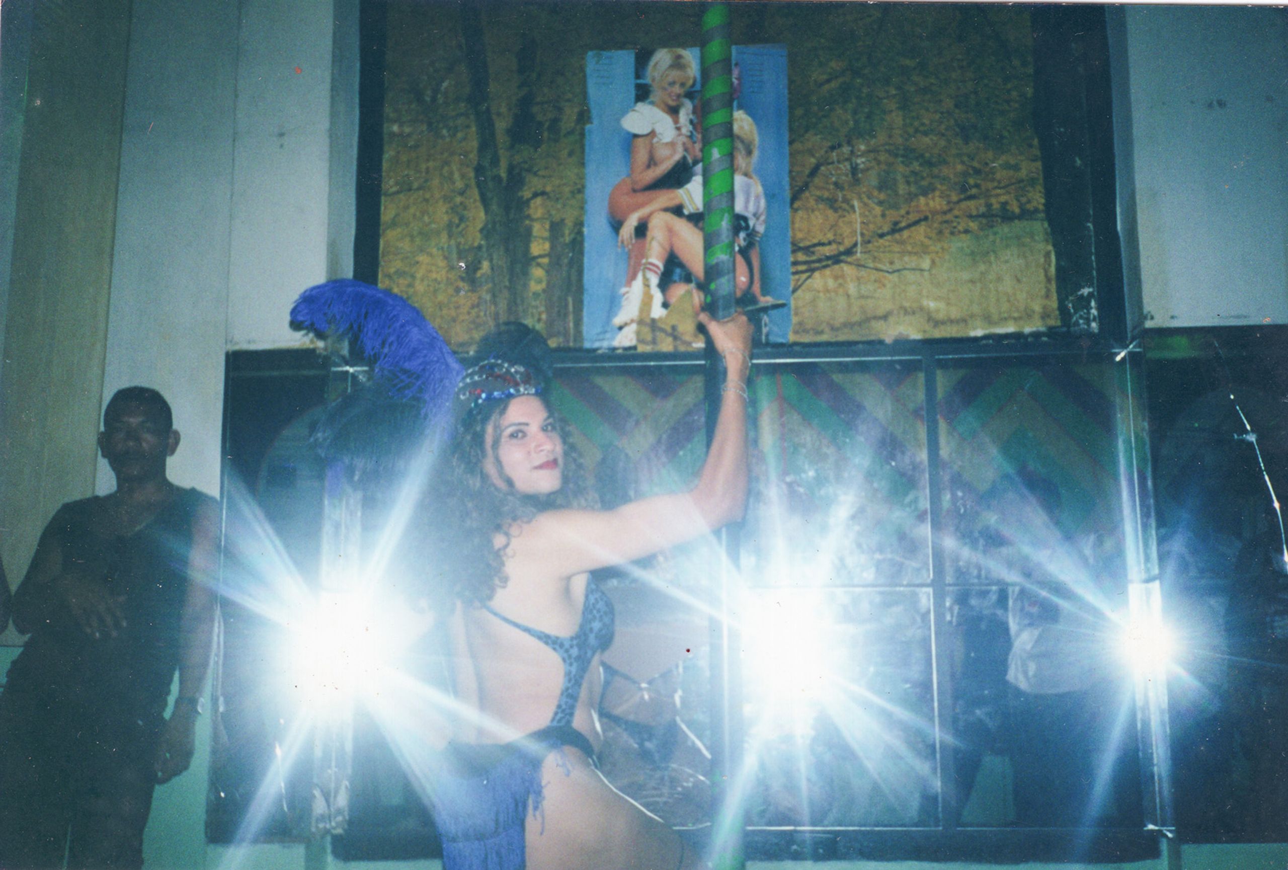 A woman in a sparkly blue costume and headpiece holds a green-striped prop, partially obscured by two bright lens flares, with a man, blue parrot statue, and posters in the background.