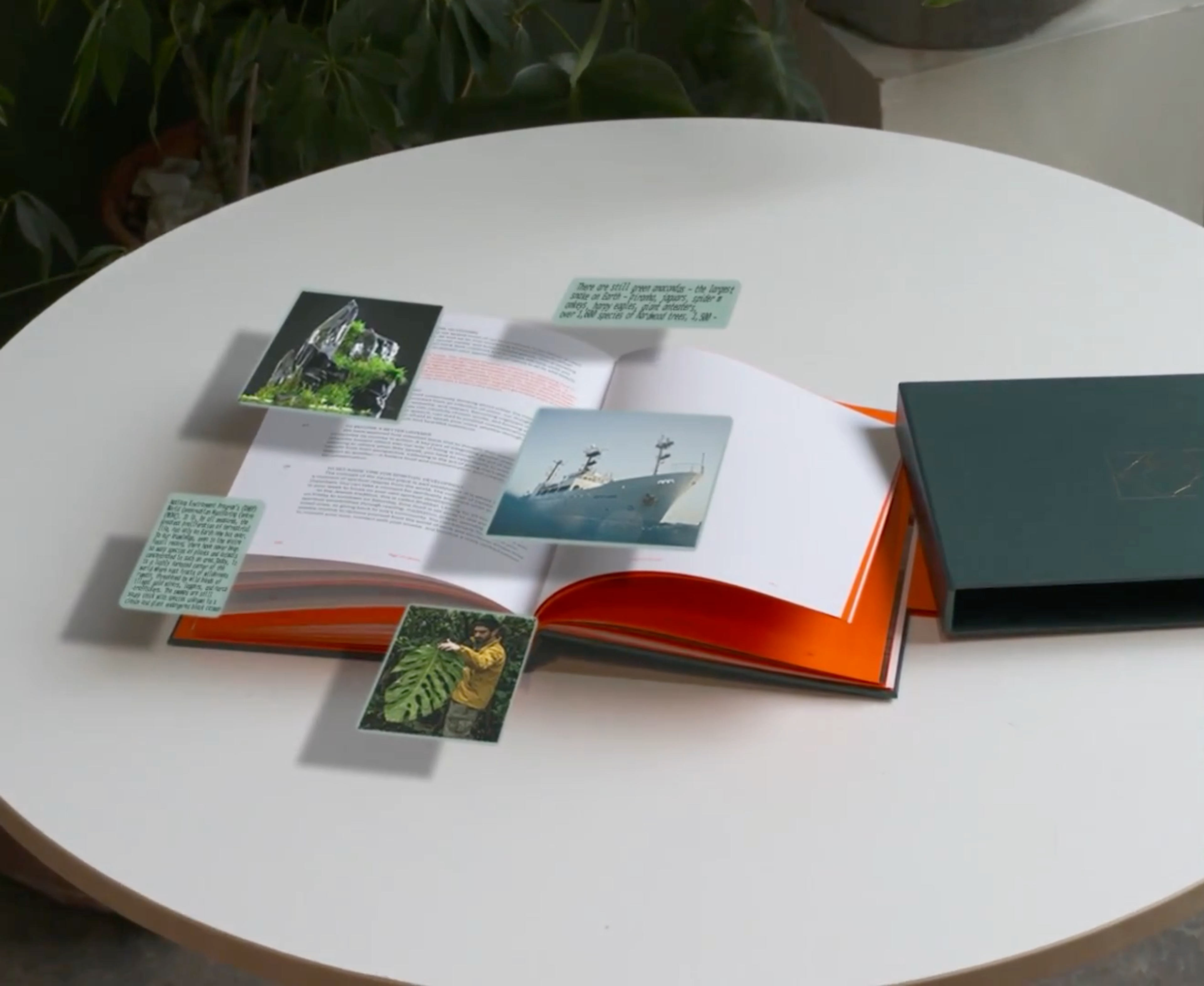An open book on a white table features digital images of a mountain, a ship, and a man with a large leaf, along with text boxes, floating above its pages. A dark green box rests next to the book.