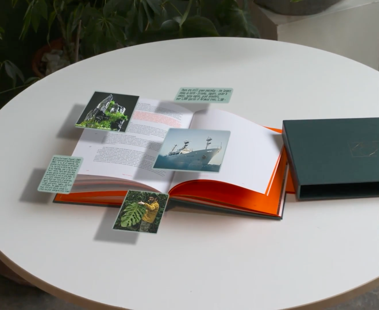 An open book on a white table features digital images of a mountain, a ship, and a man with a large leaf, along with text boxes, floating above its pages. A dark green box rests next to the book.
