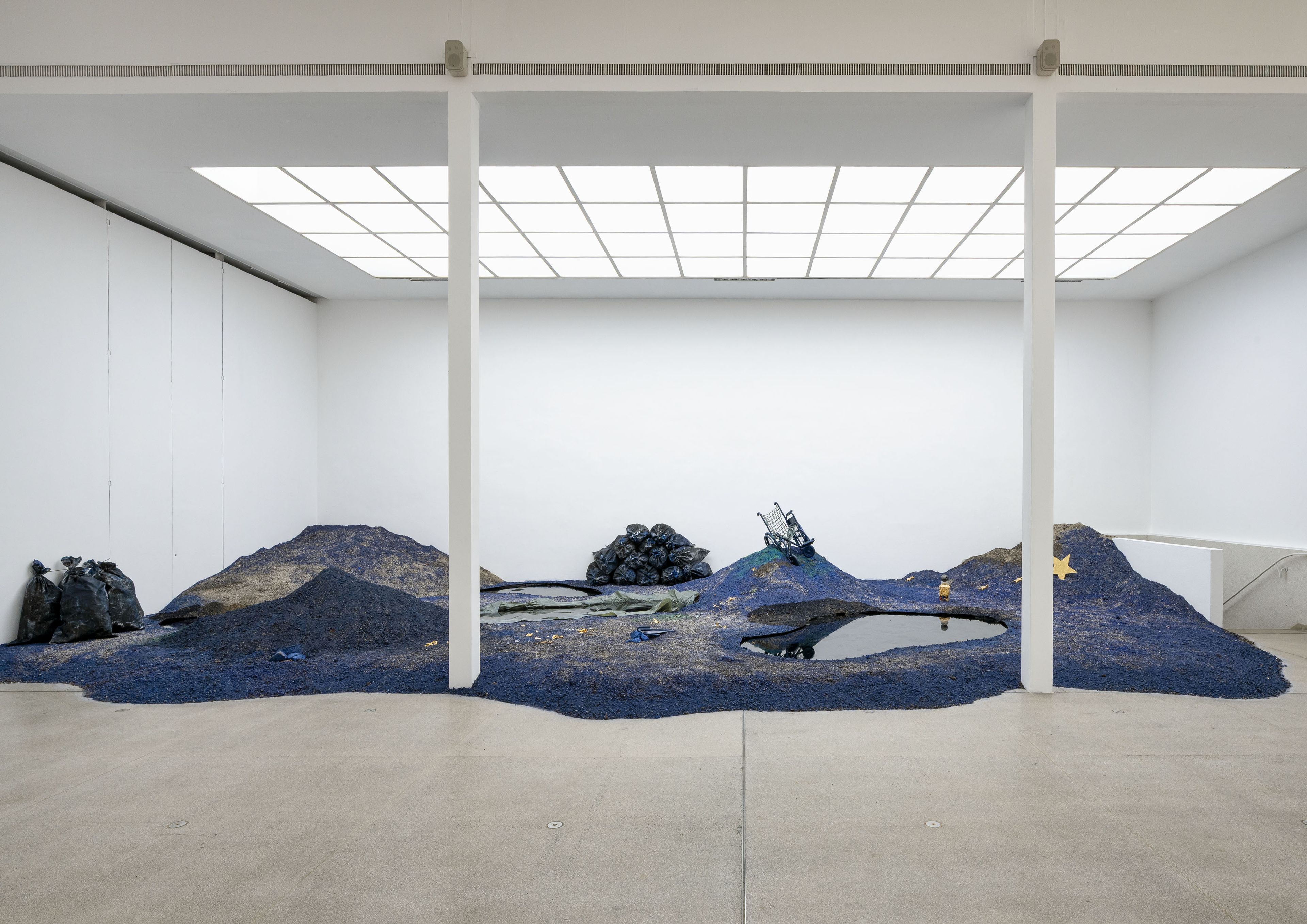 Art installation of a dark blue and brown landscape with black trash bags, debris, and a reflective pool in a white gallery.
