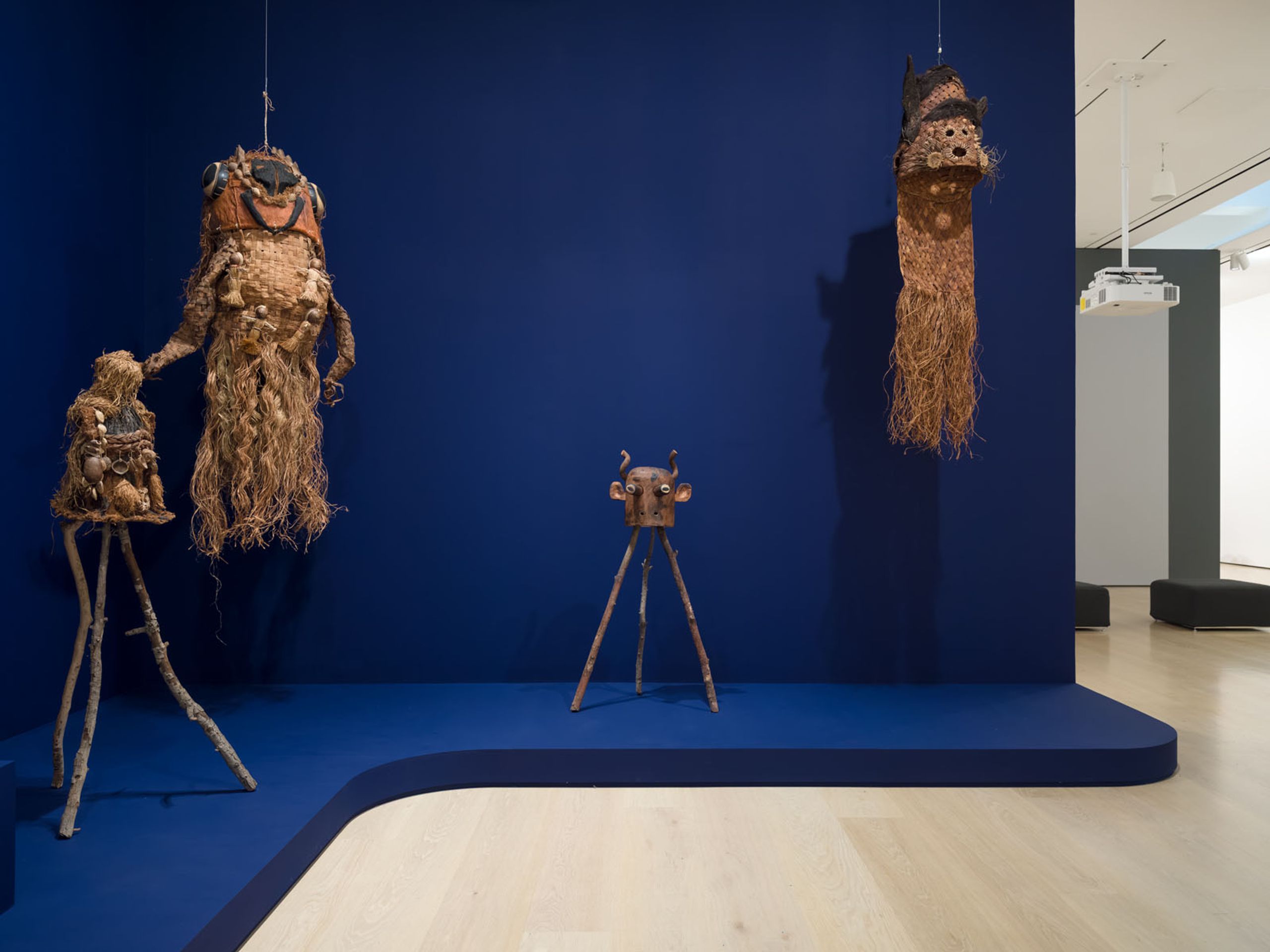 An art gallery installation featuring three organic fiber sculptures and a metallic bull's head, set against a deep blue wall and floor.