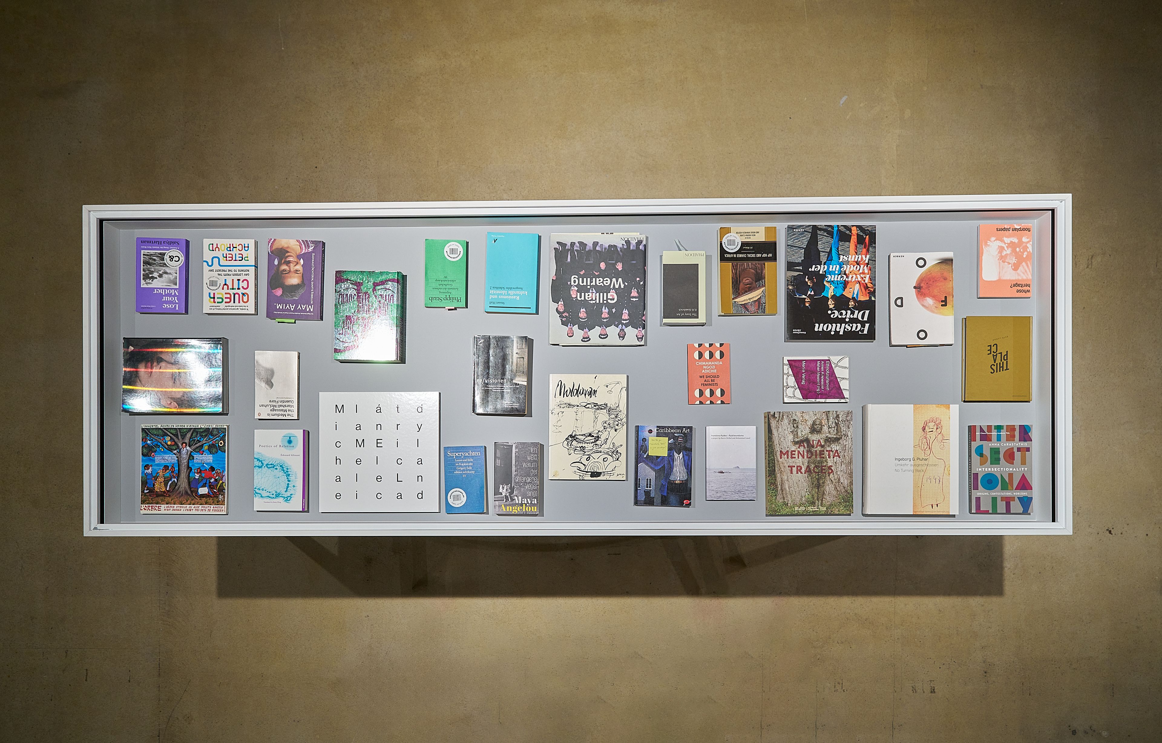 A white-framed display case filled with numerous small books and zines, mounted on a concrete wall.