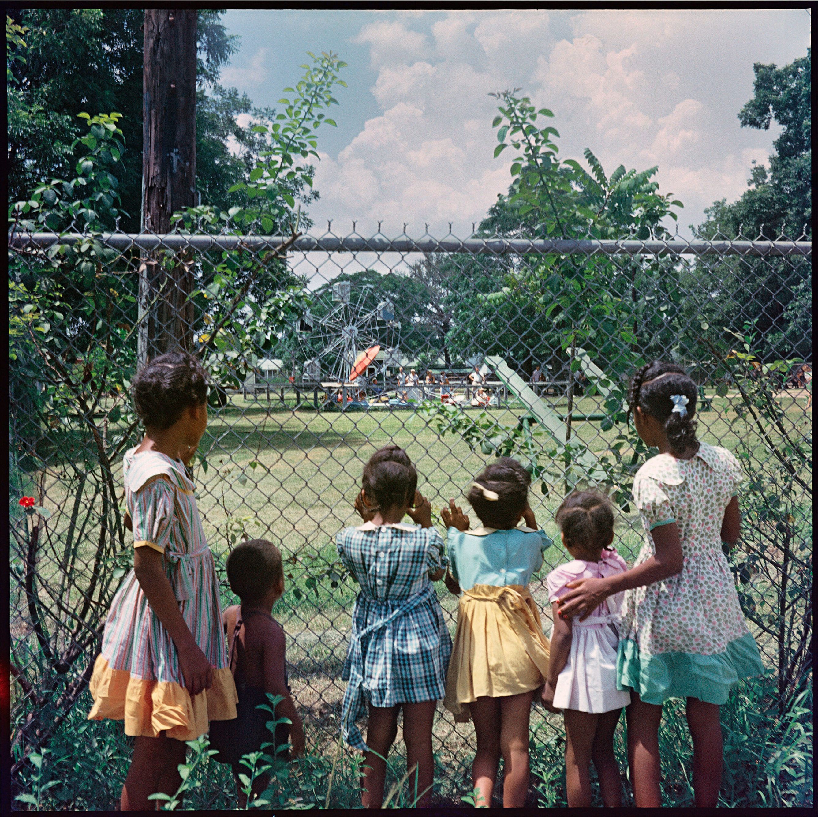 Six young Black children gaze through a chain-link fence at a distant amusement park with a Ferris wheel.