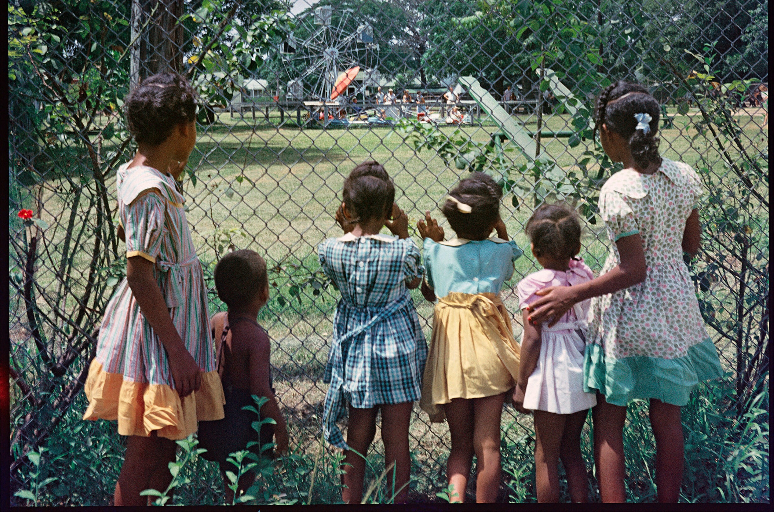Six young Black children gaze through a chain-link fence at a distant amusement park with a Ferris wheel.