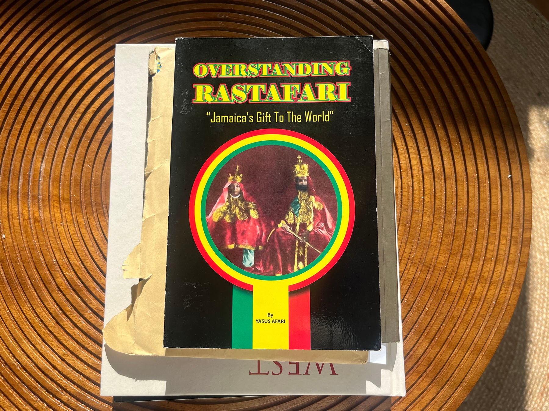 The cover of the book "OVERSTANDING RASTAFARI" featuring two crowned, regal figures within a red, yellow, and green circle, resting on other books.