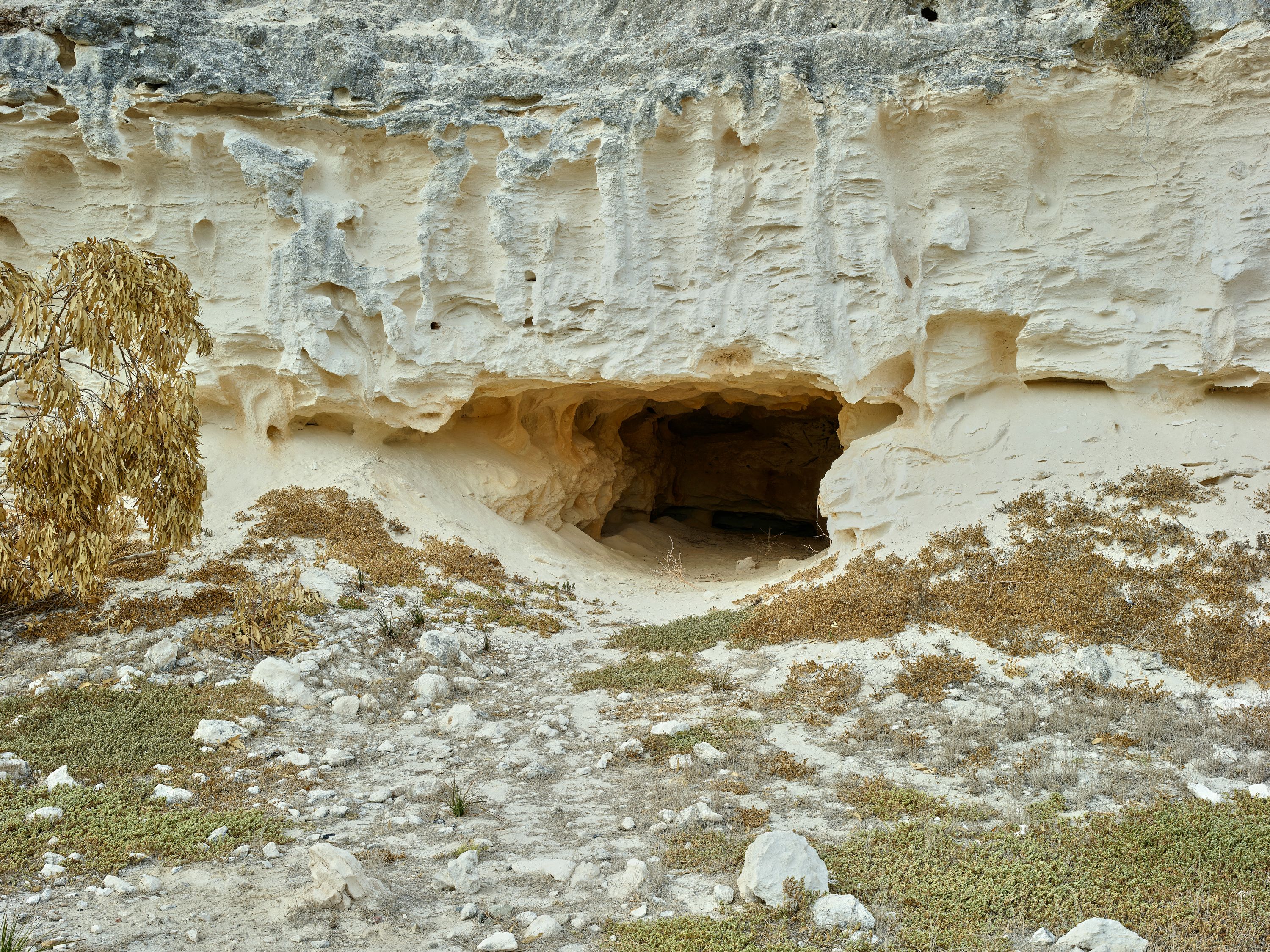 A dark cave entrance in a light-colored, eroded rock cliff with sparse vegetation.