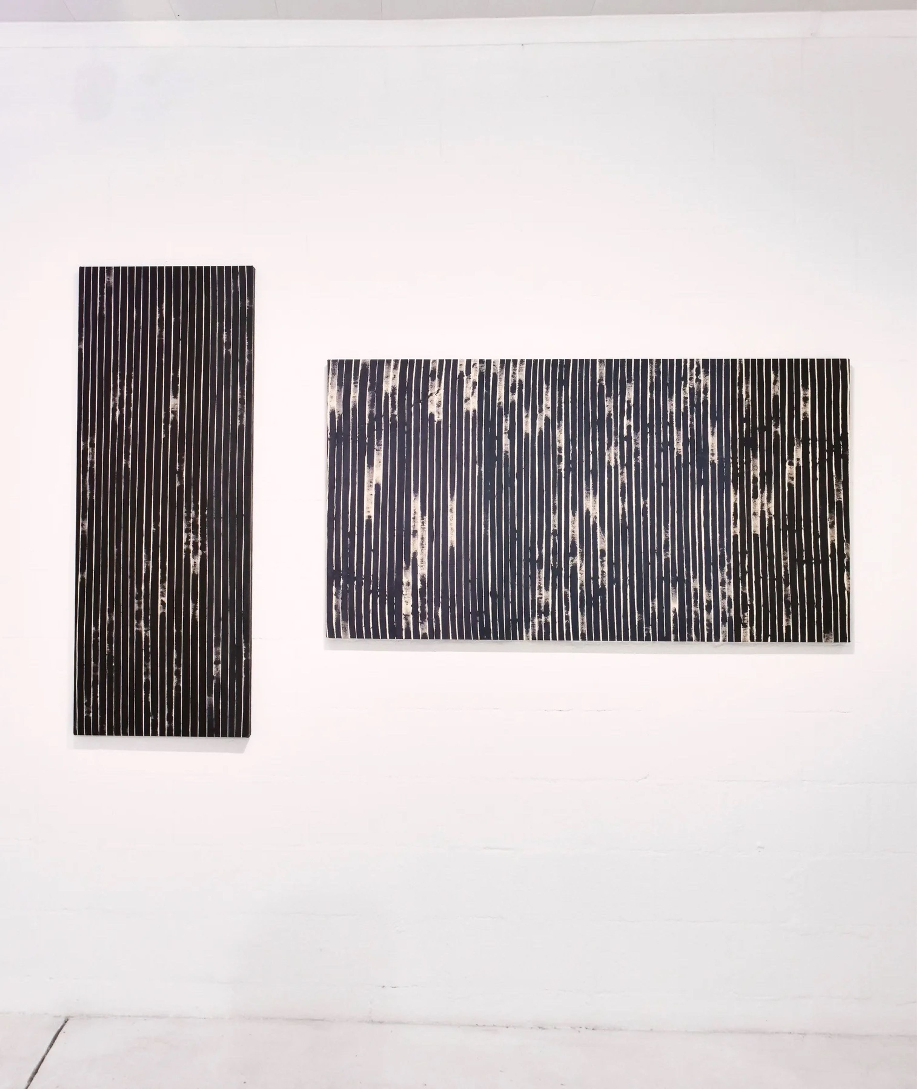 Two abstract paintings on a white wall, one vertical and one horizontal, both with dark, textured vertical stripes against a lighter background.