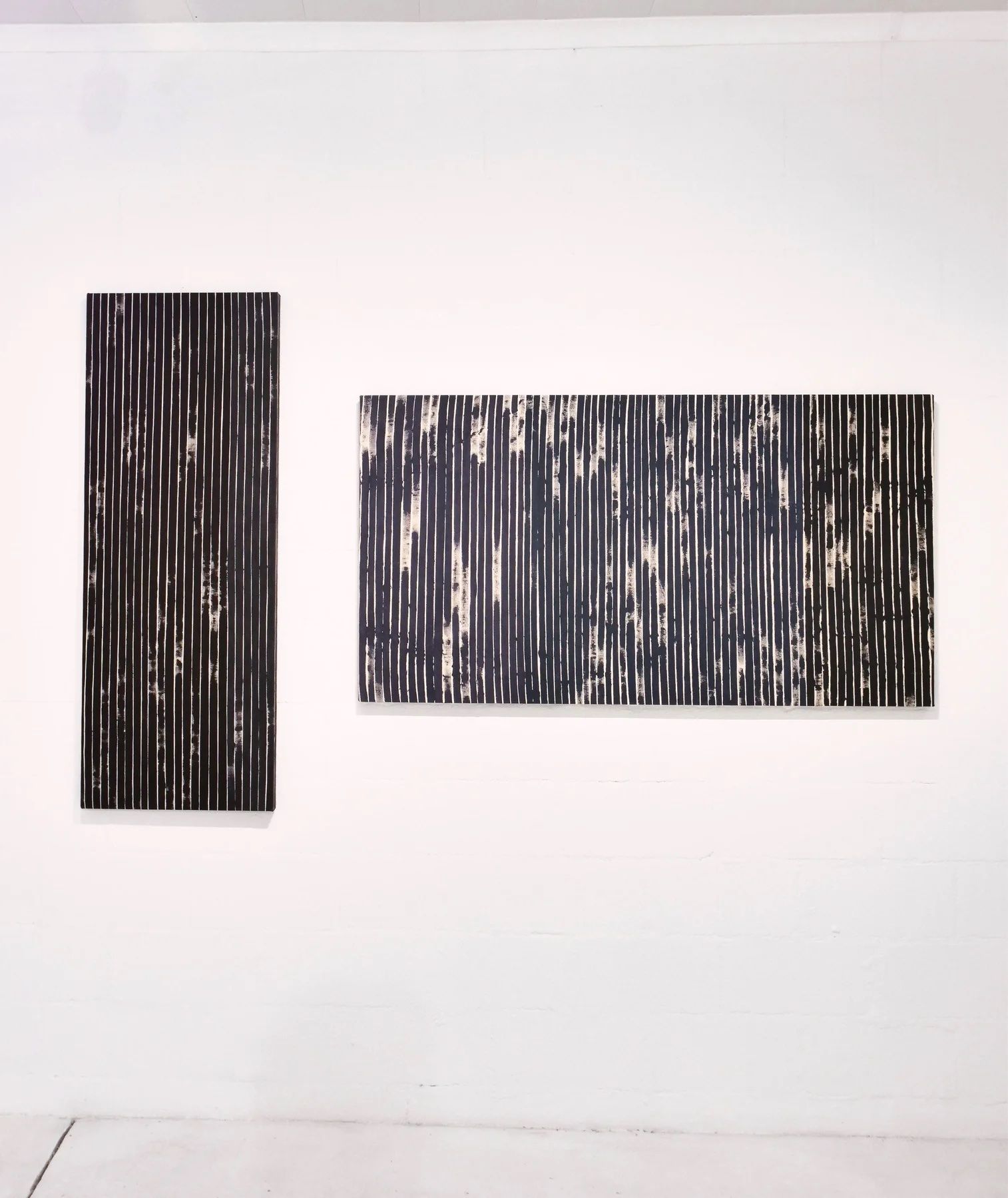 Two abstract paintings on a white wall, one vertical and one horizontal, both with dark, textured vertical stripes against a lighter background.