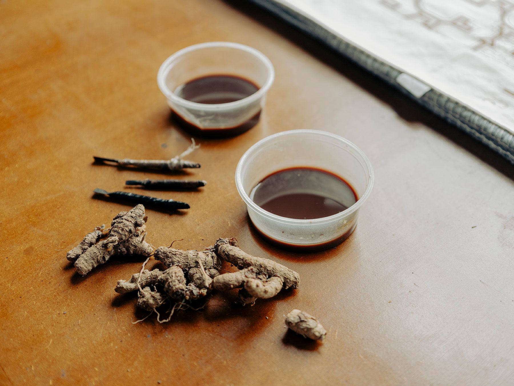 Two small cups of dark liquid, a pile of dry gnarled roots, and a few dark stick-like objects on a brown surface.