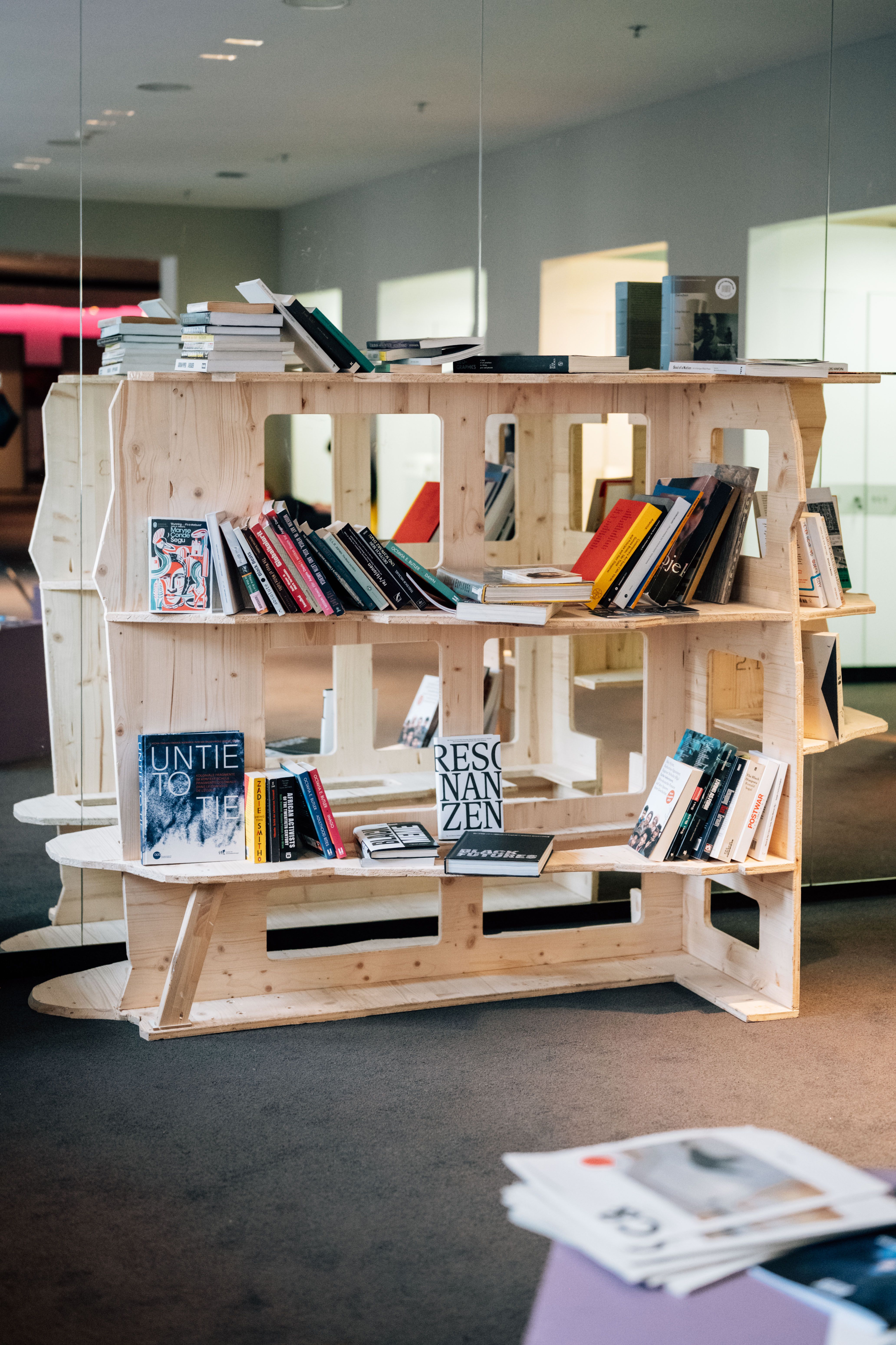 A modular light wood bookshelf holds numerous books in a bright room with reflective walls.