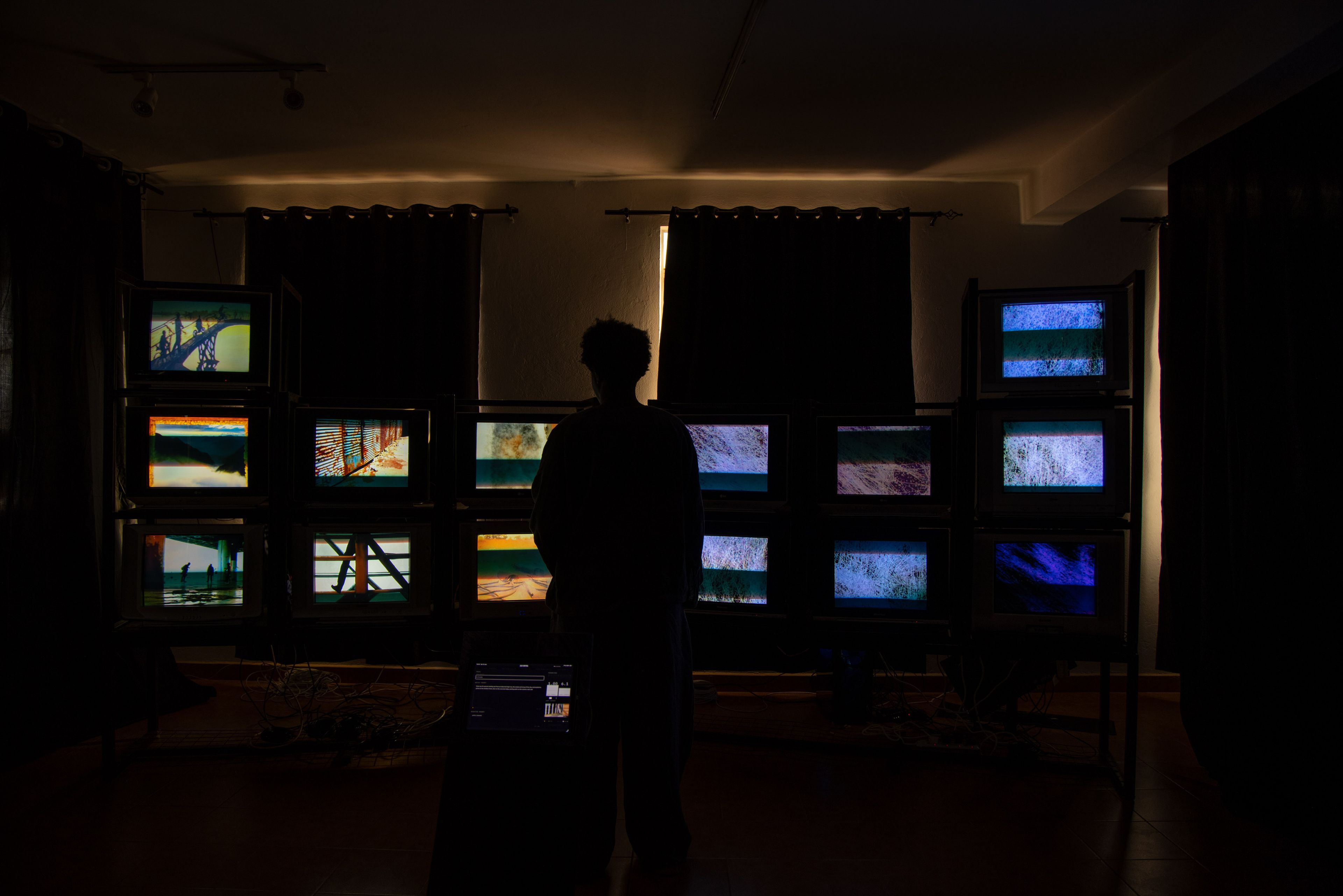 A person silhouetted in a dark room faces a wall of multiple CRT televisions displaying various images and static.