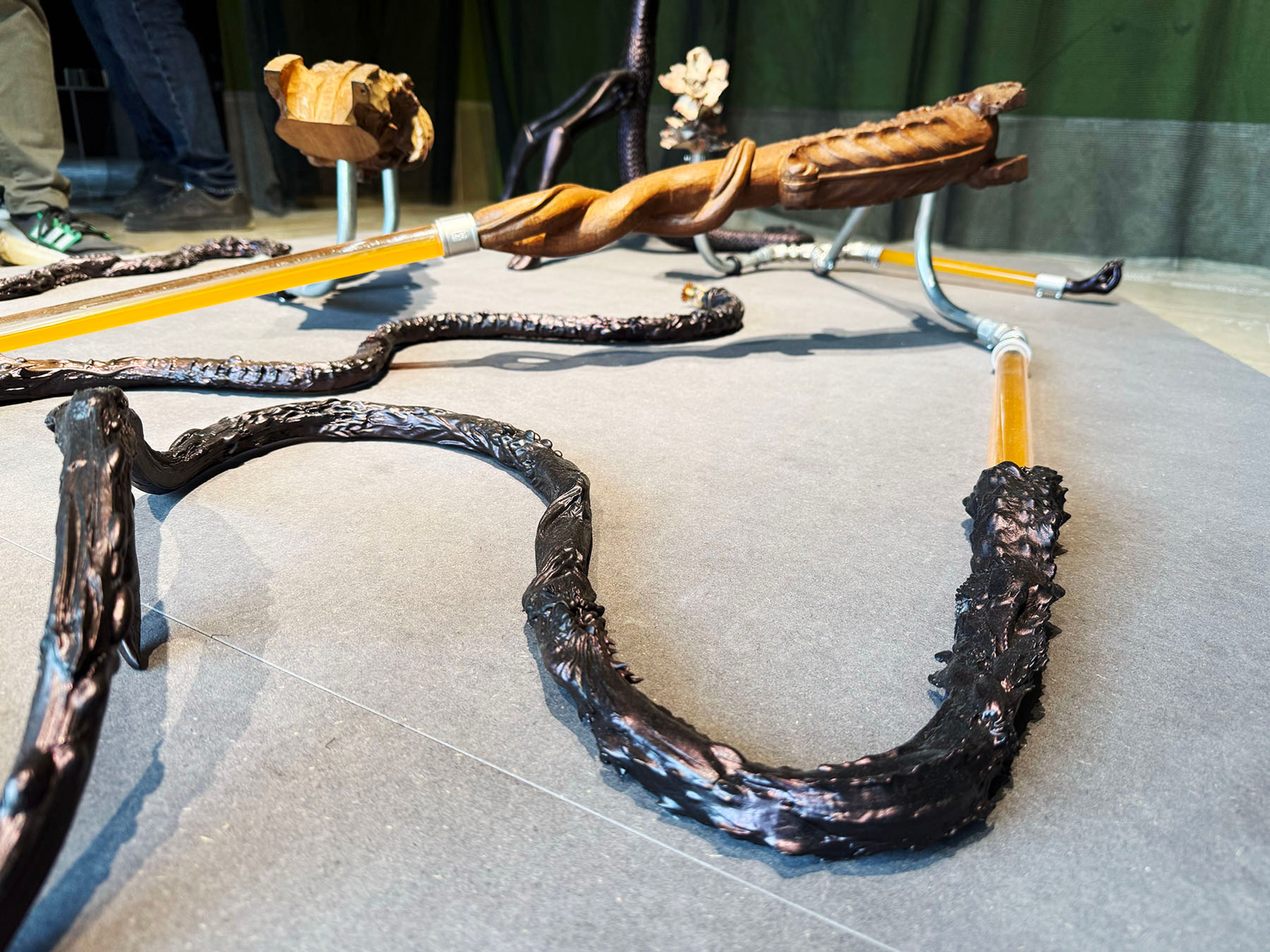 An art installation of dark, gnarled organic forms connected by translucent amber tubes and metal fittings on a grey floor.