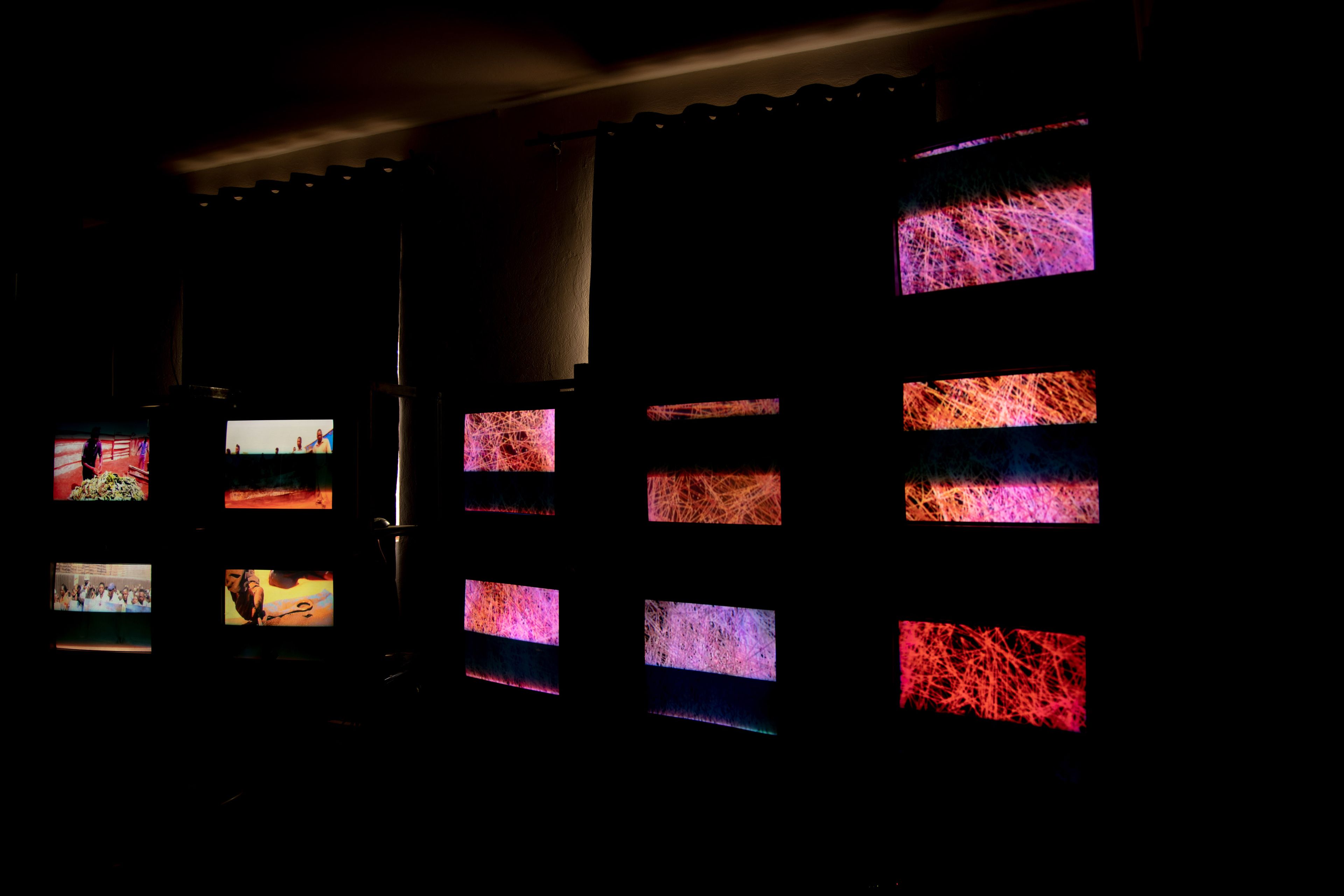 Multiple screens in a dark room. Some screens on the left show various scenes, while screens on the right display abstract patterns of red, orange, and pink lines.
