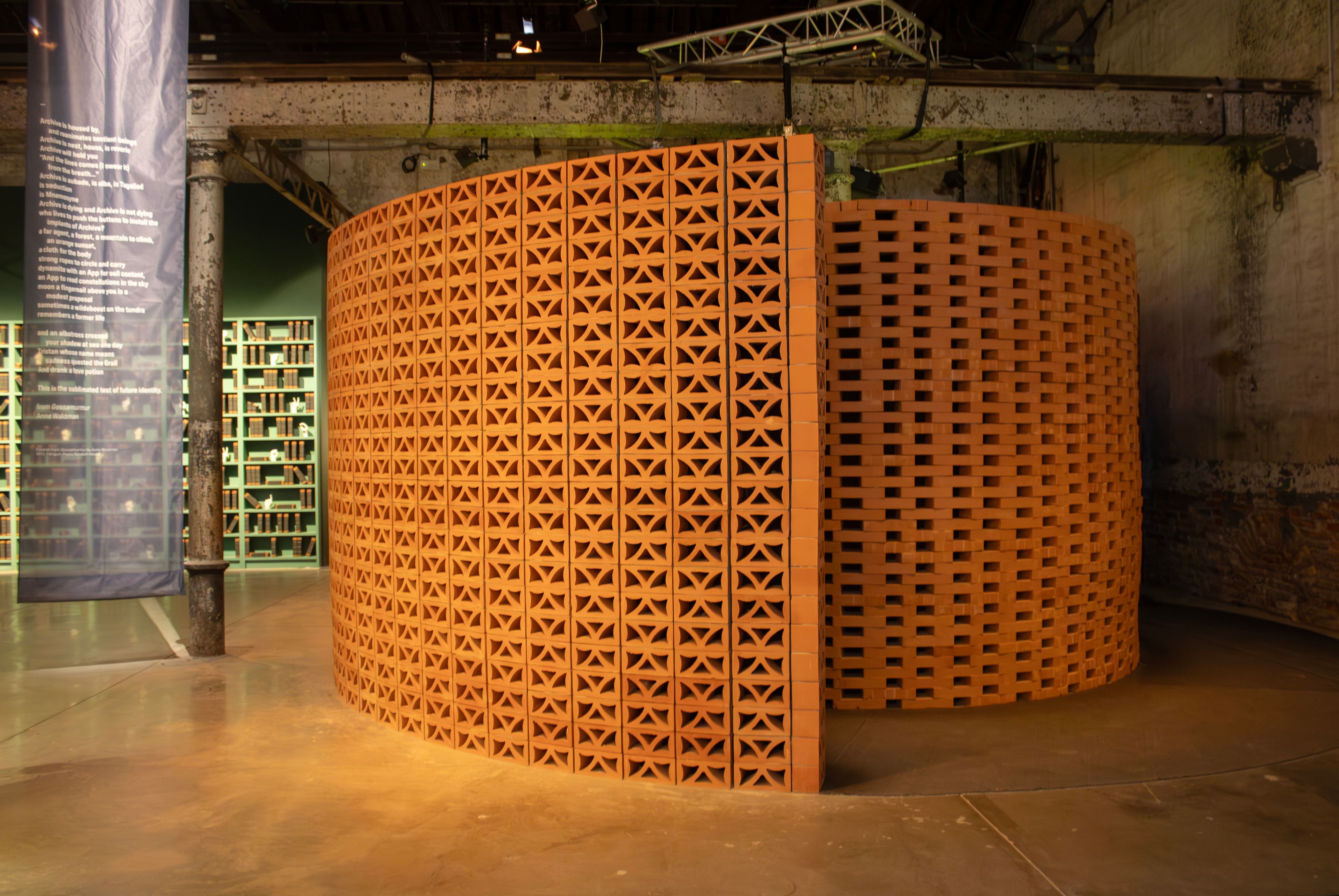 A curved wall of reddish-brown perforated bricks with geometric patterns stands in a dimly lit exhibition space.