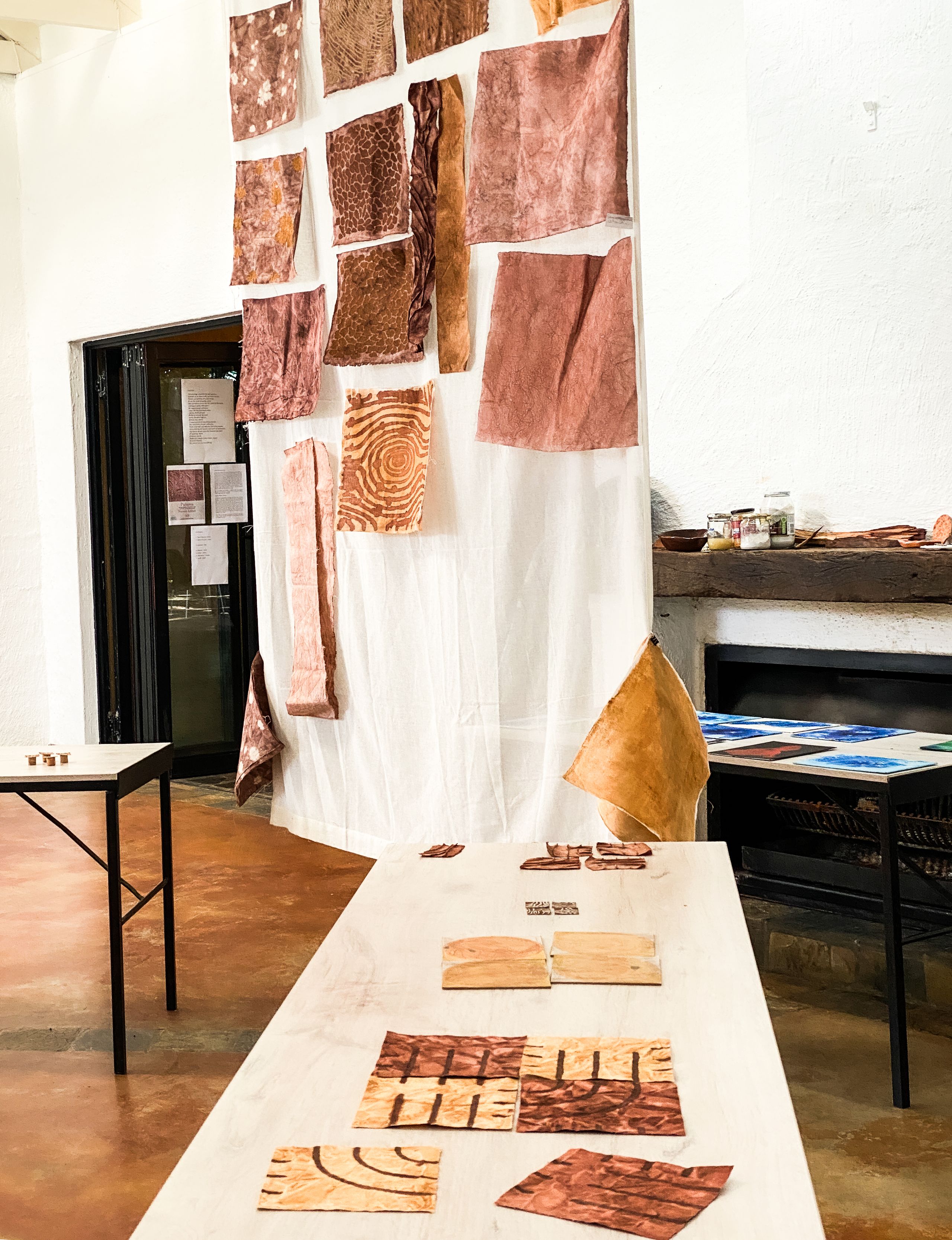 An art exhibition featuring various patterned, earthy-toned fabric or bark pieces hanging on a white wall and smaller samples laid on a long table.