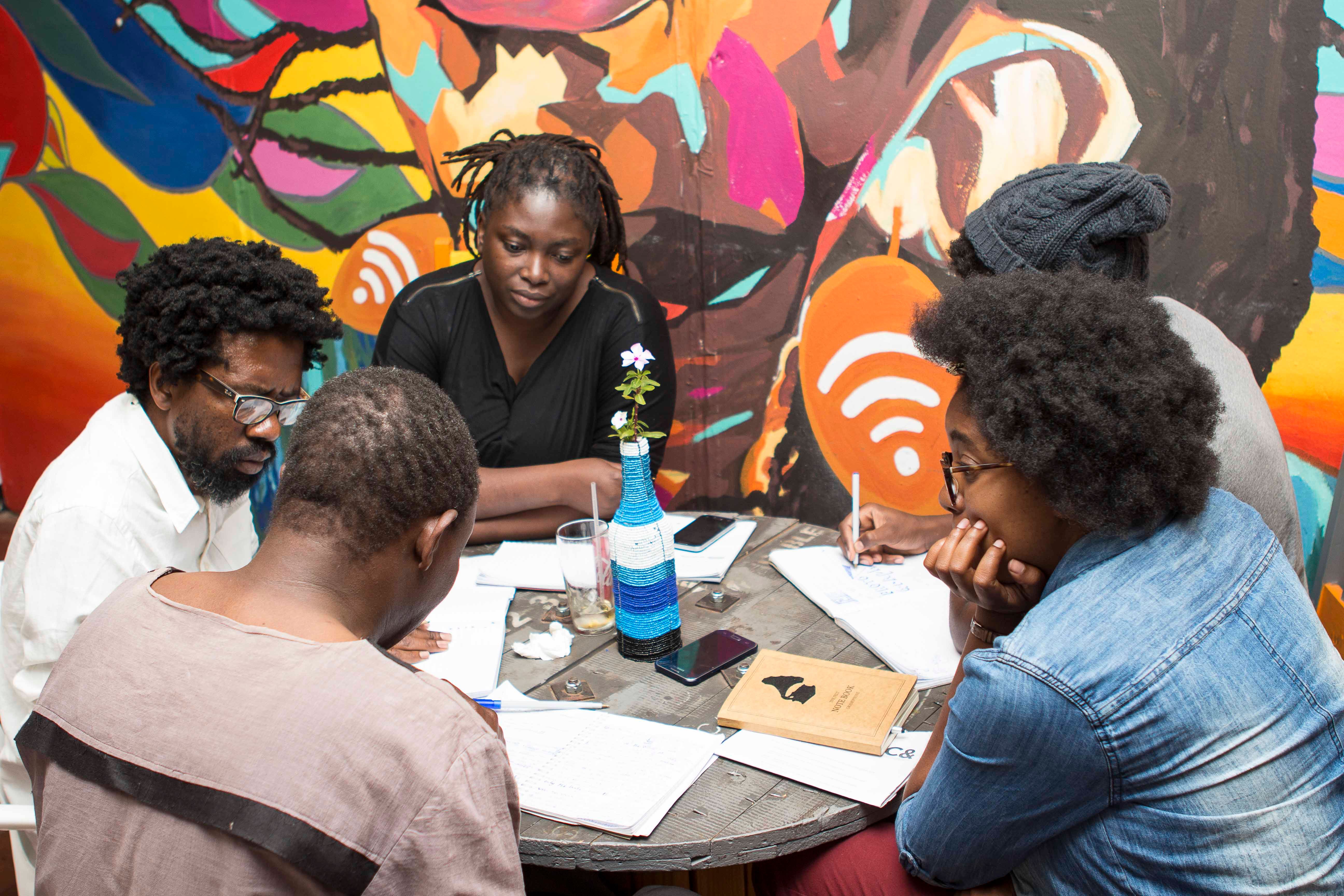 Five Black people gathered around a table, writing and discussing, against a vibrant mural backdrop.