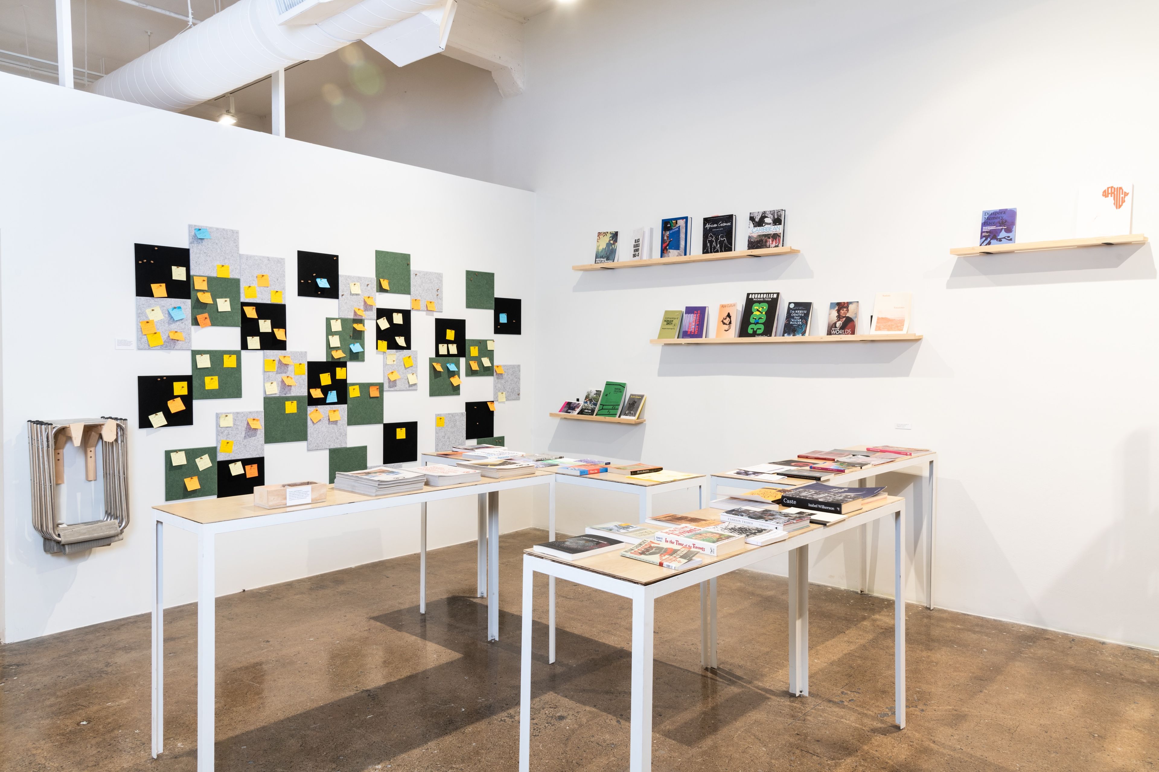 A modern gallery space with a wall display of sticky notes on square panels, and tables and shelves filled with books and magazines.