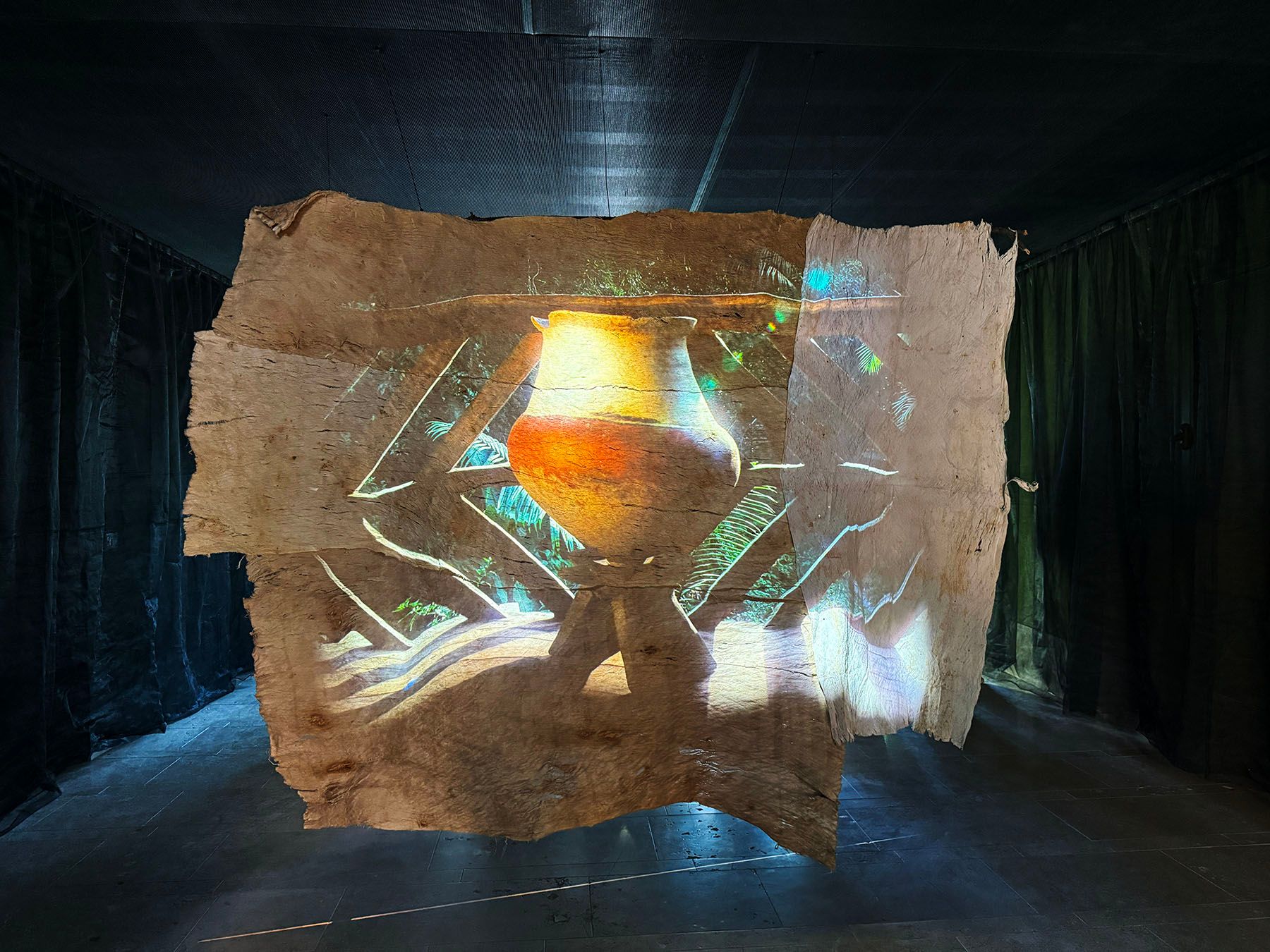 A projection of a clay pot on a stand is cast onto an irregular, bark-like sheet in a dark room.