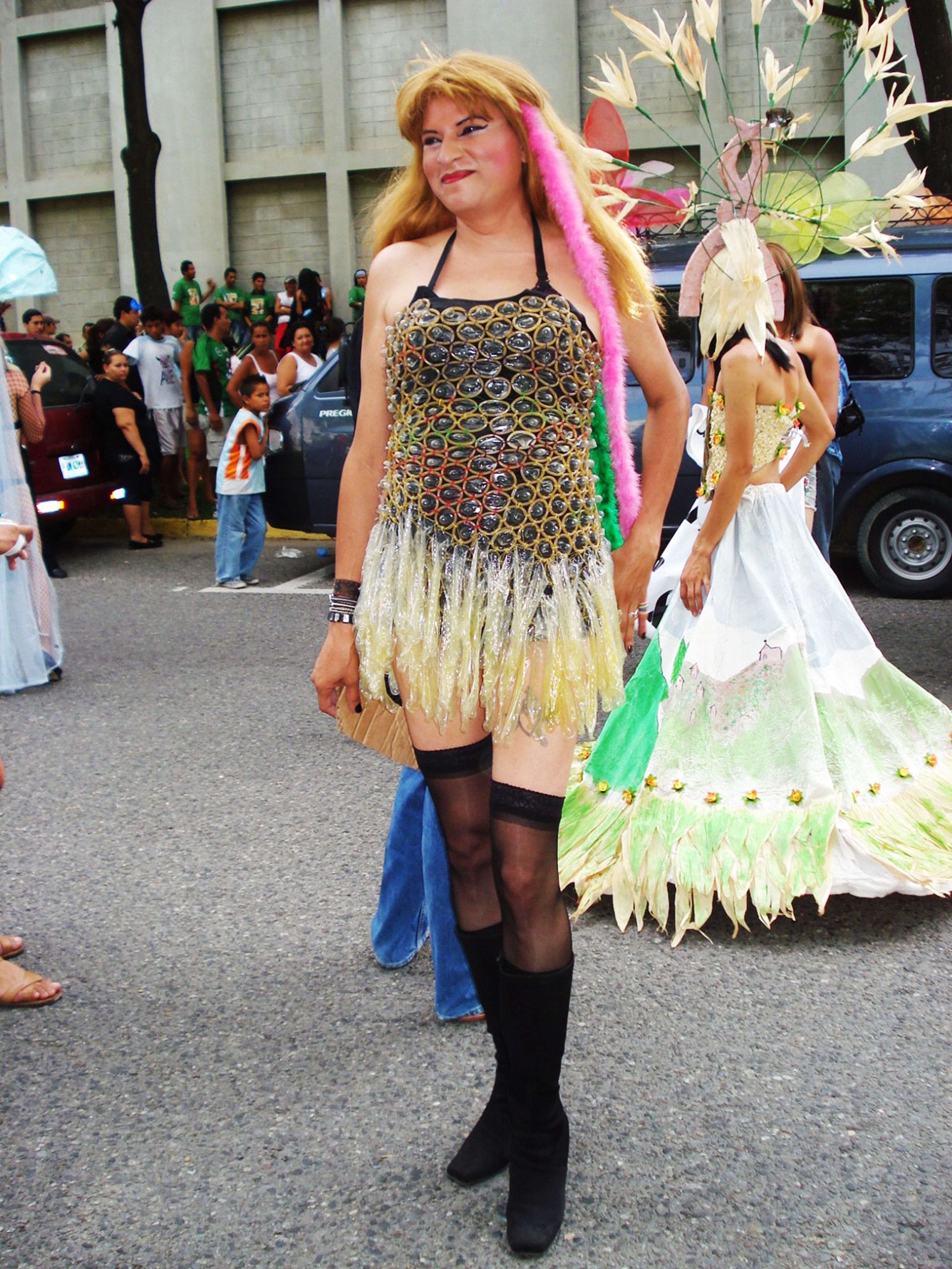 A person in a flamboyant dress made of condom wrappers with a feathered boa, thigh-high stockings, and boots, smiling at a parade.