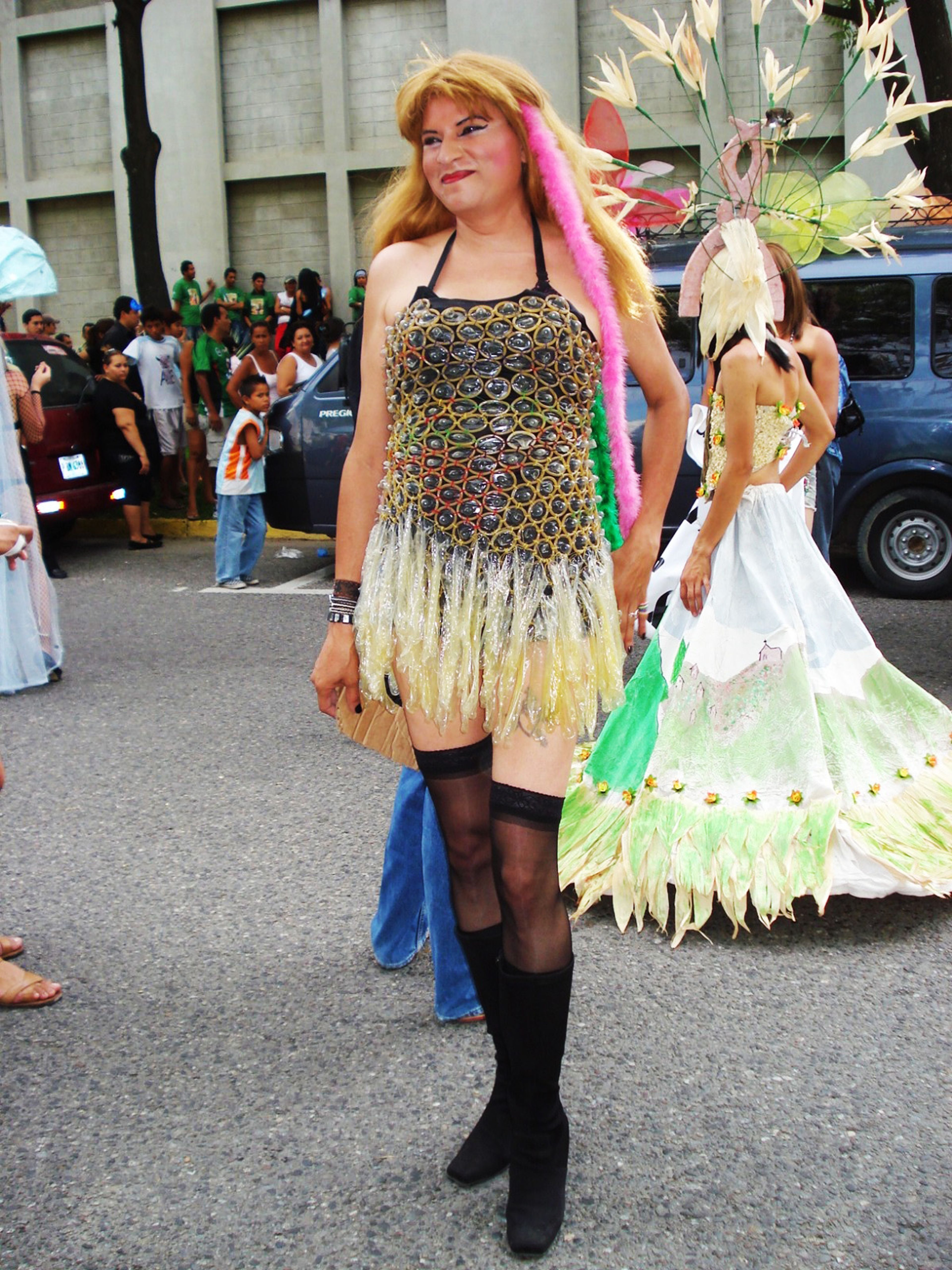 A person in a flamboyant dress made of condom wrappers with a feathered boa, thigh-high stockings, and boots, smiling at a parade.