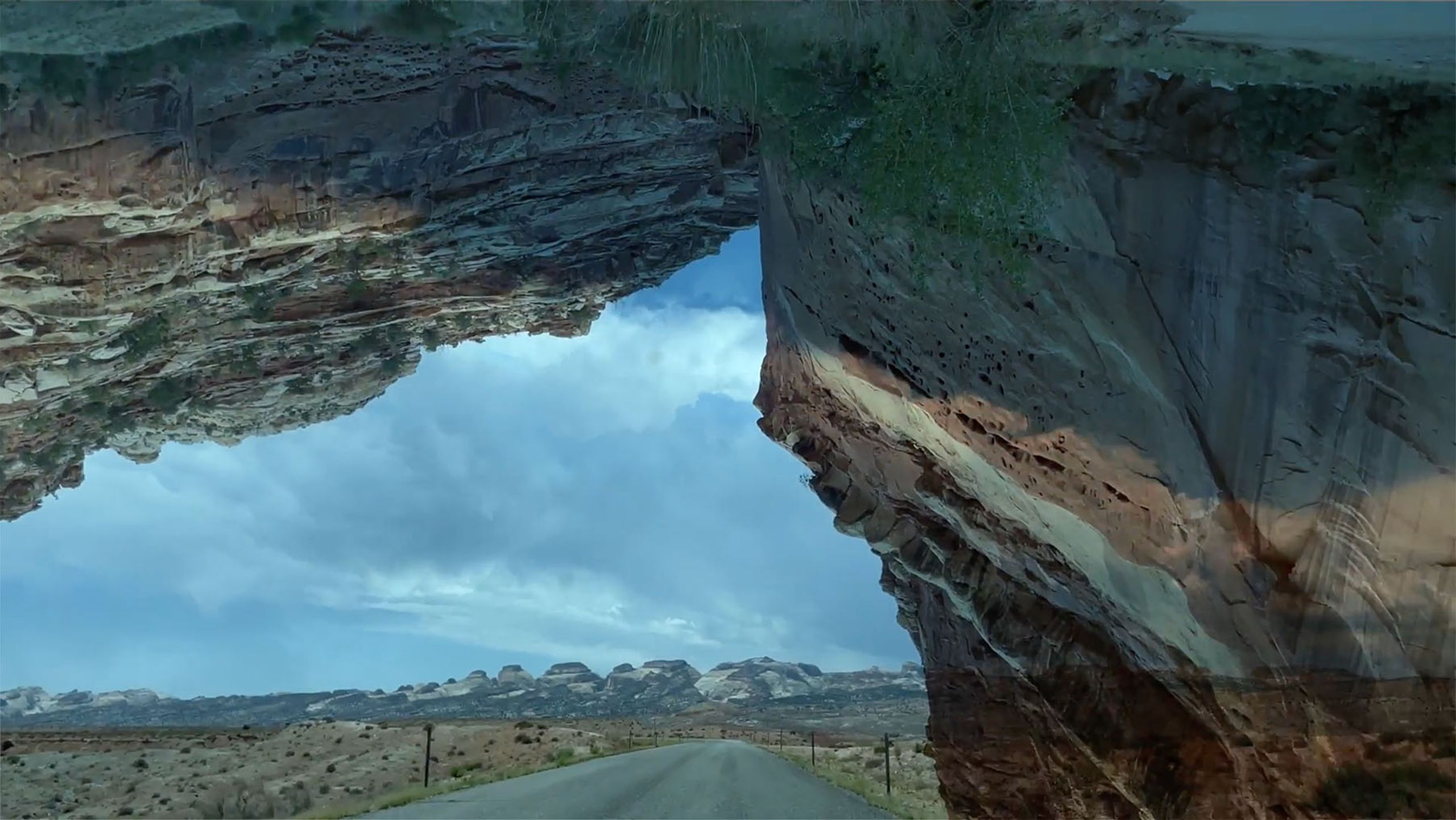 A road passes under a natural rock arch into a desert landscape with distant mesas under a cloudy sky.