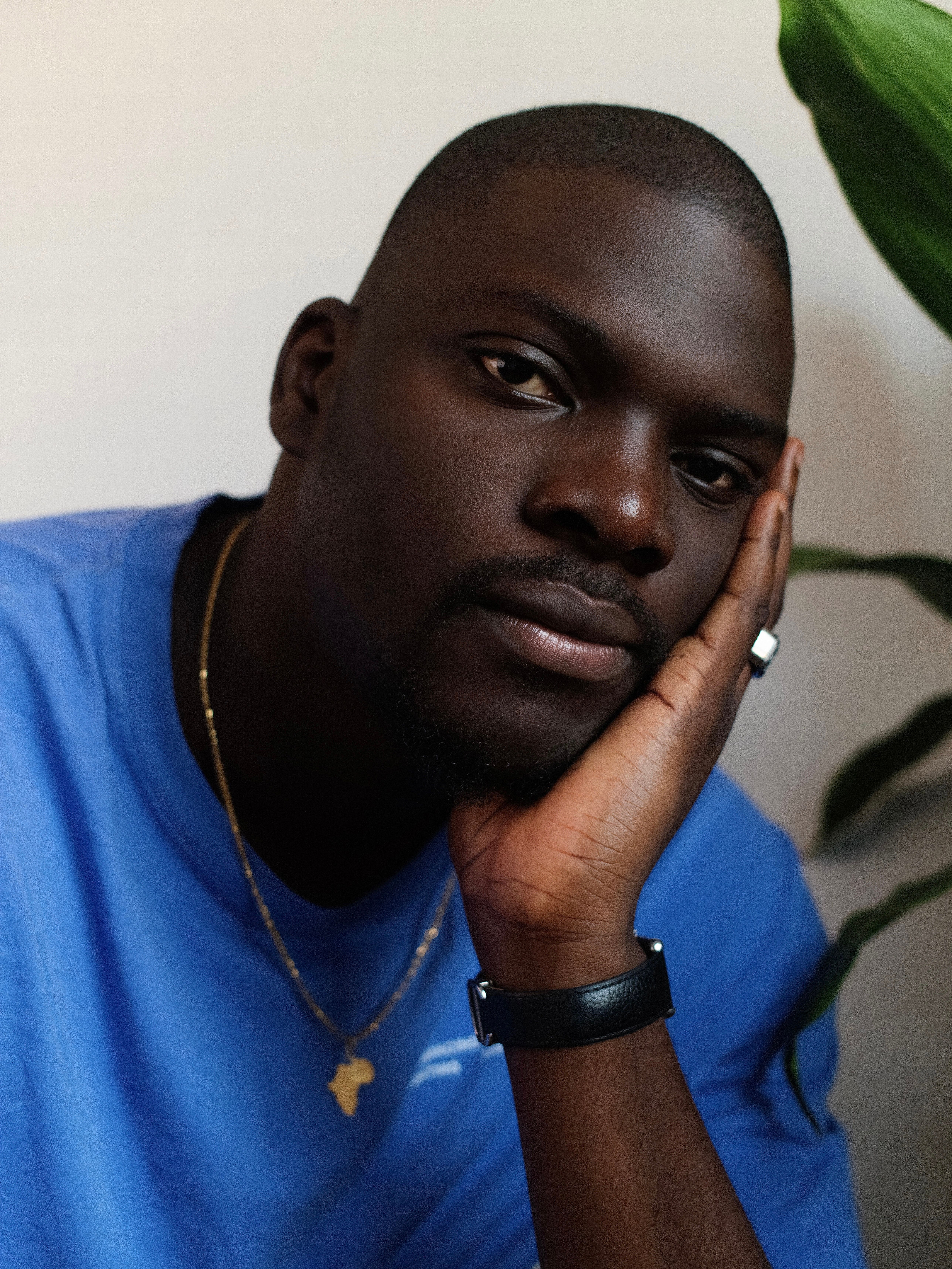 A dark-skinned man in a blue shirt, wearing an Africa pendant, rests his hand on his chin and looks at the camera.
