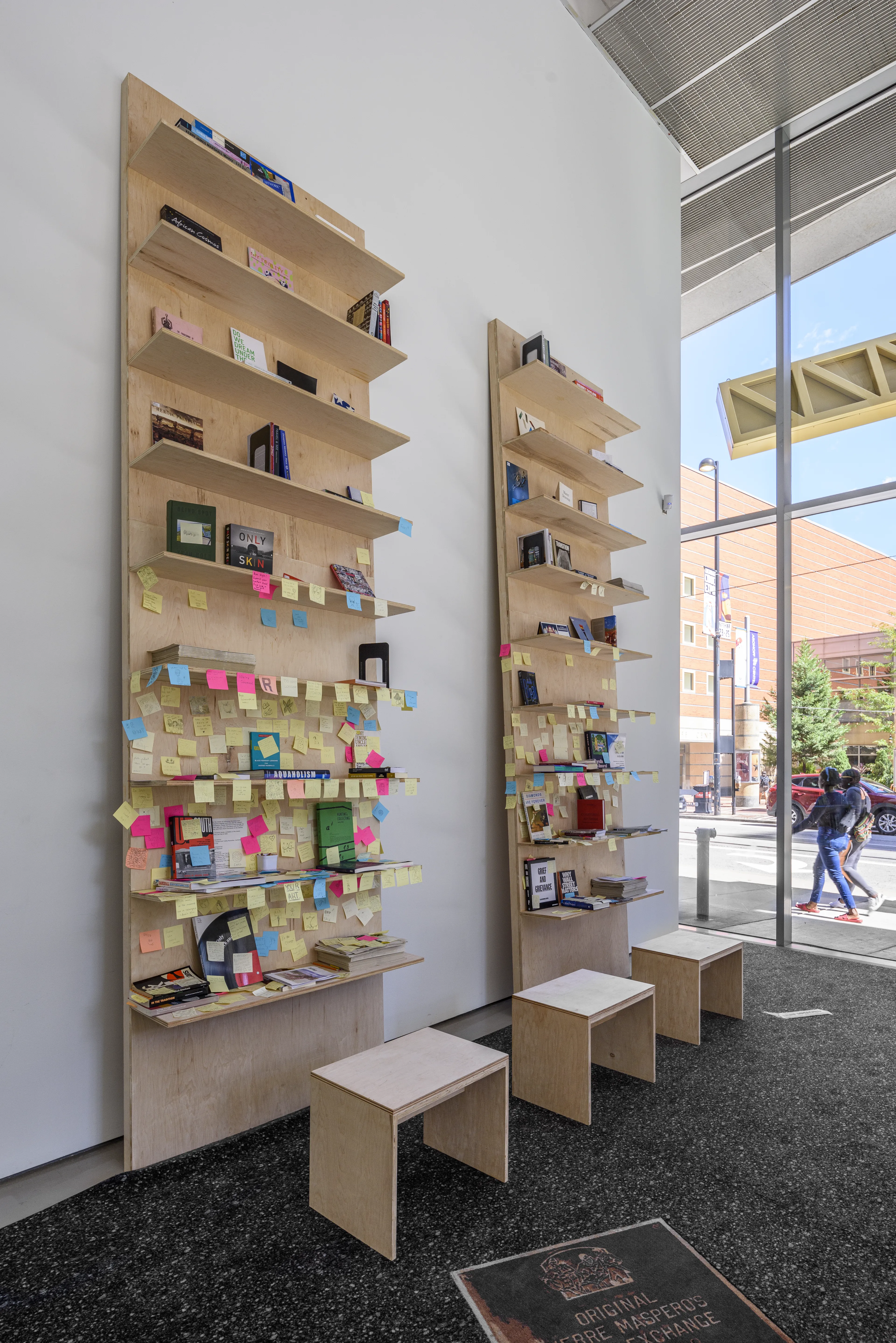 Two tall wooden bookshelves filled with books and colorful sticky notes stand in a bright room with large windows overlooking a city street, with three small wooden stools in the foreground.