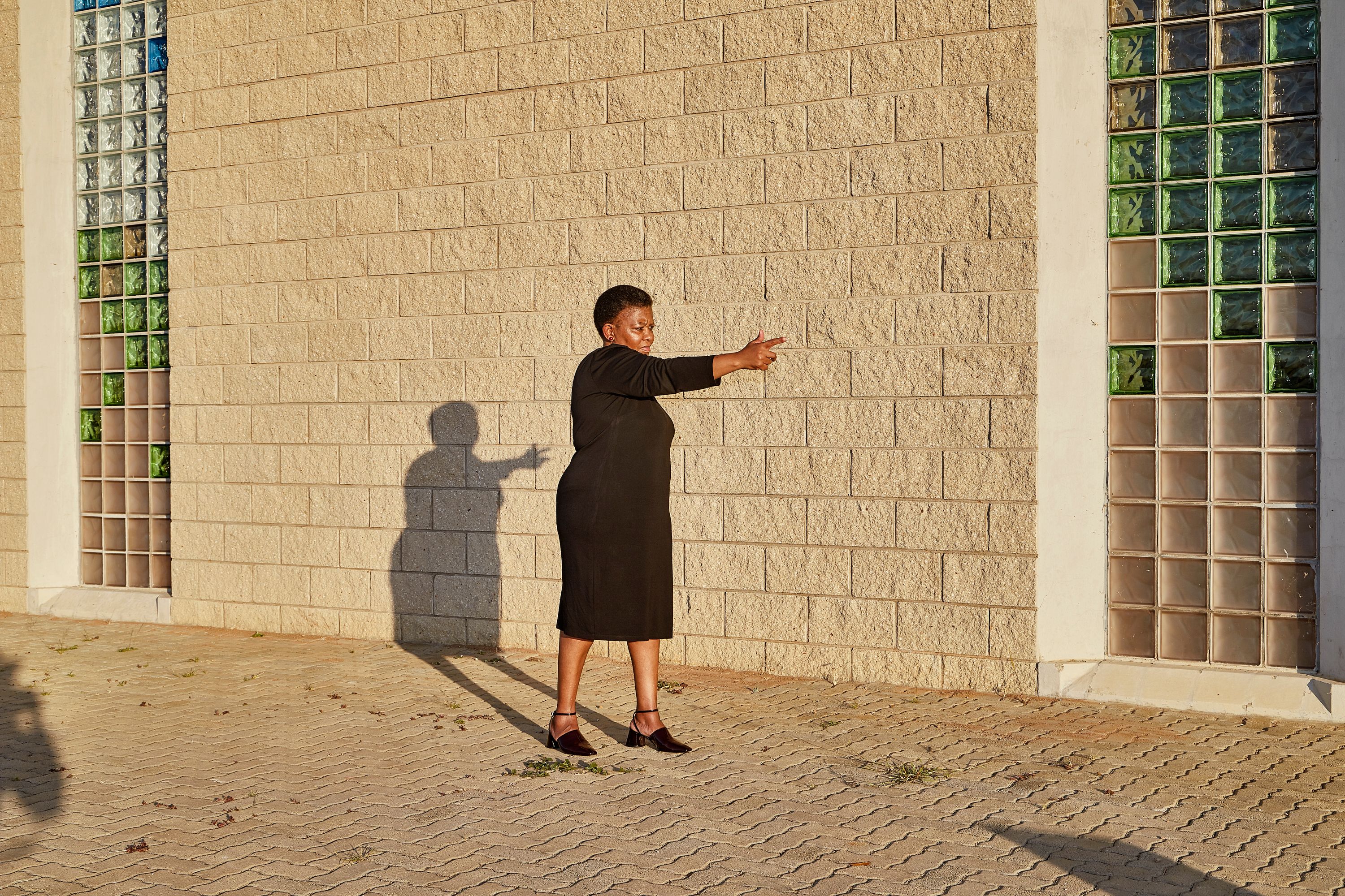 A Black woman in a black dress stands against a brick wall, pointing right, with a long shadow.