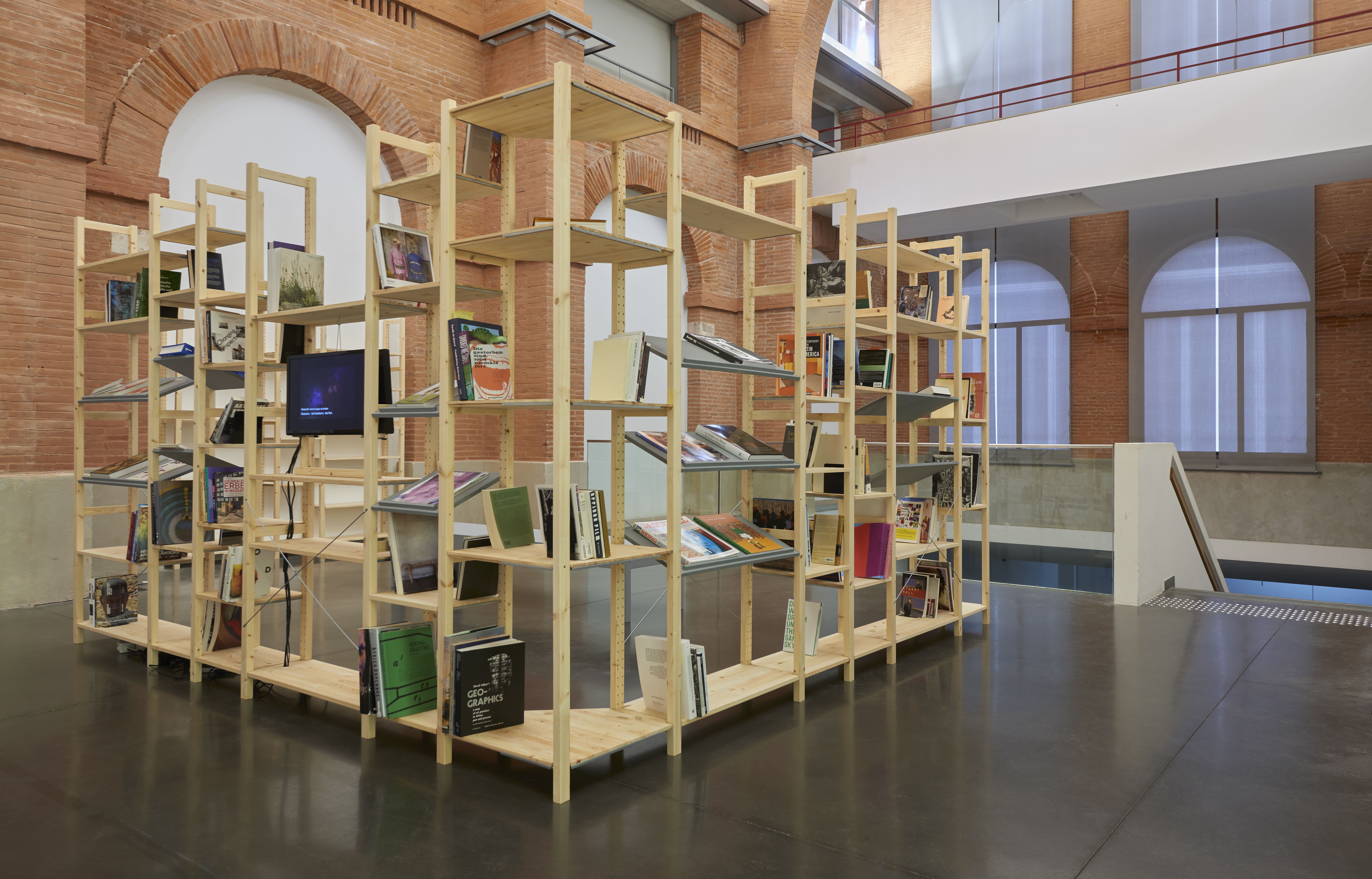 L-shaped wooden shelving unit displaying books and a monitor in a spacious room with brick walls.