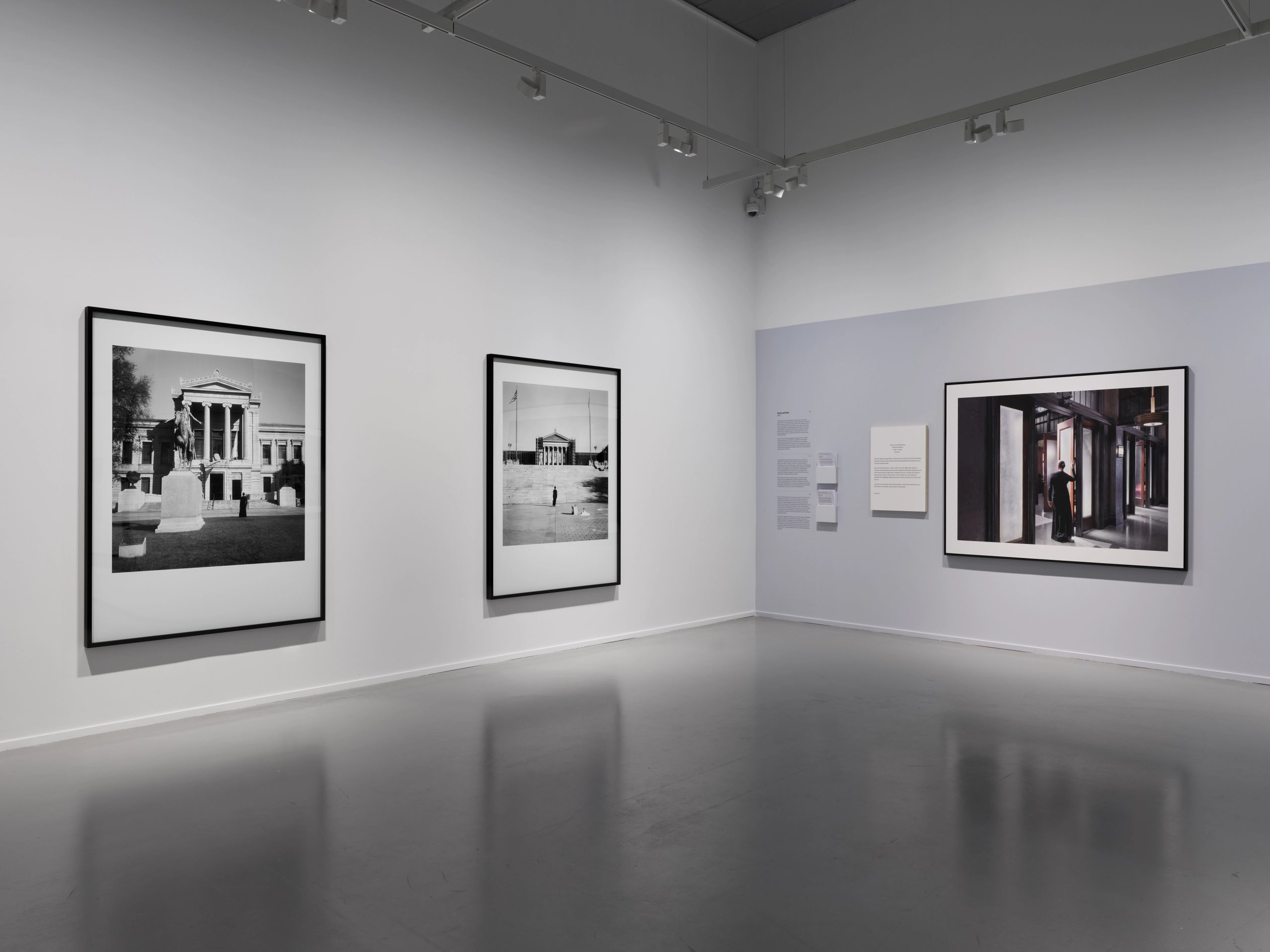 Three large framed photographs hang on white and light blue-grey walls in a modern gallery with a reflective grey floor.