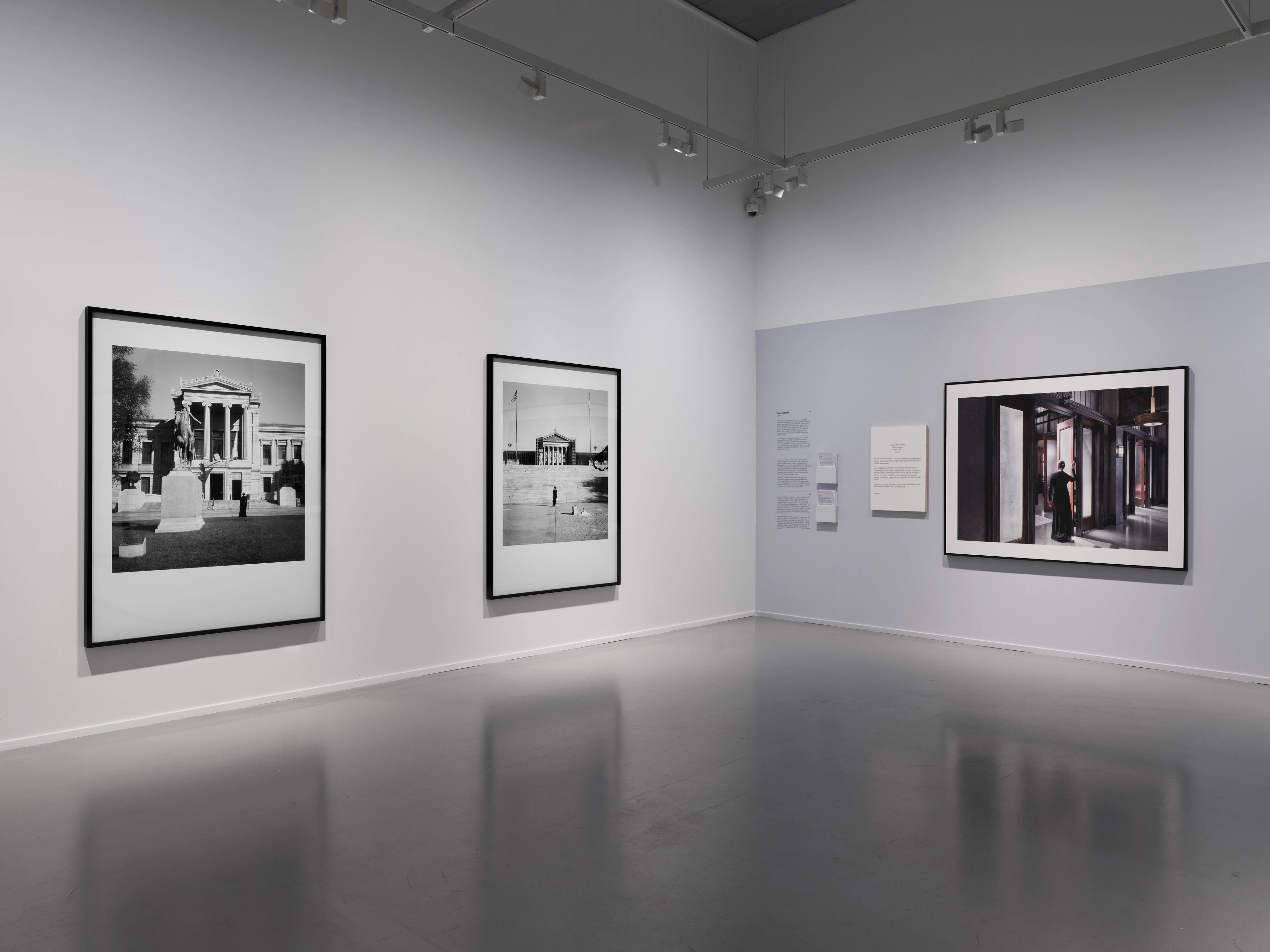 Three large framed photographs hang on white and light blue-grey walls in a modern gallery with a reflective grey floor.