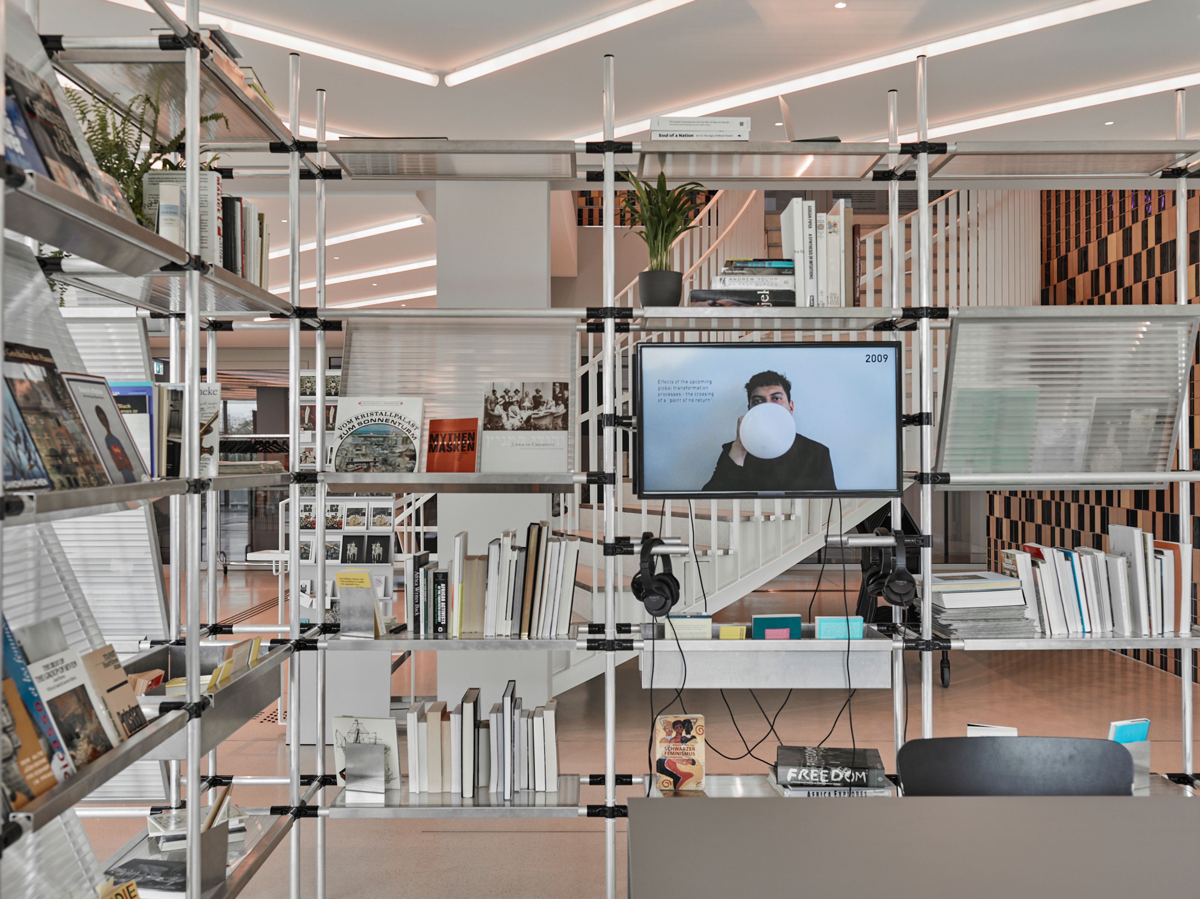 A modern metal shelving unit filled with books and a TV displaying a man holding a white balloon, in a bright, contemporary space.