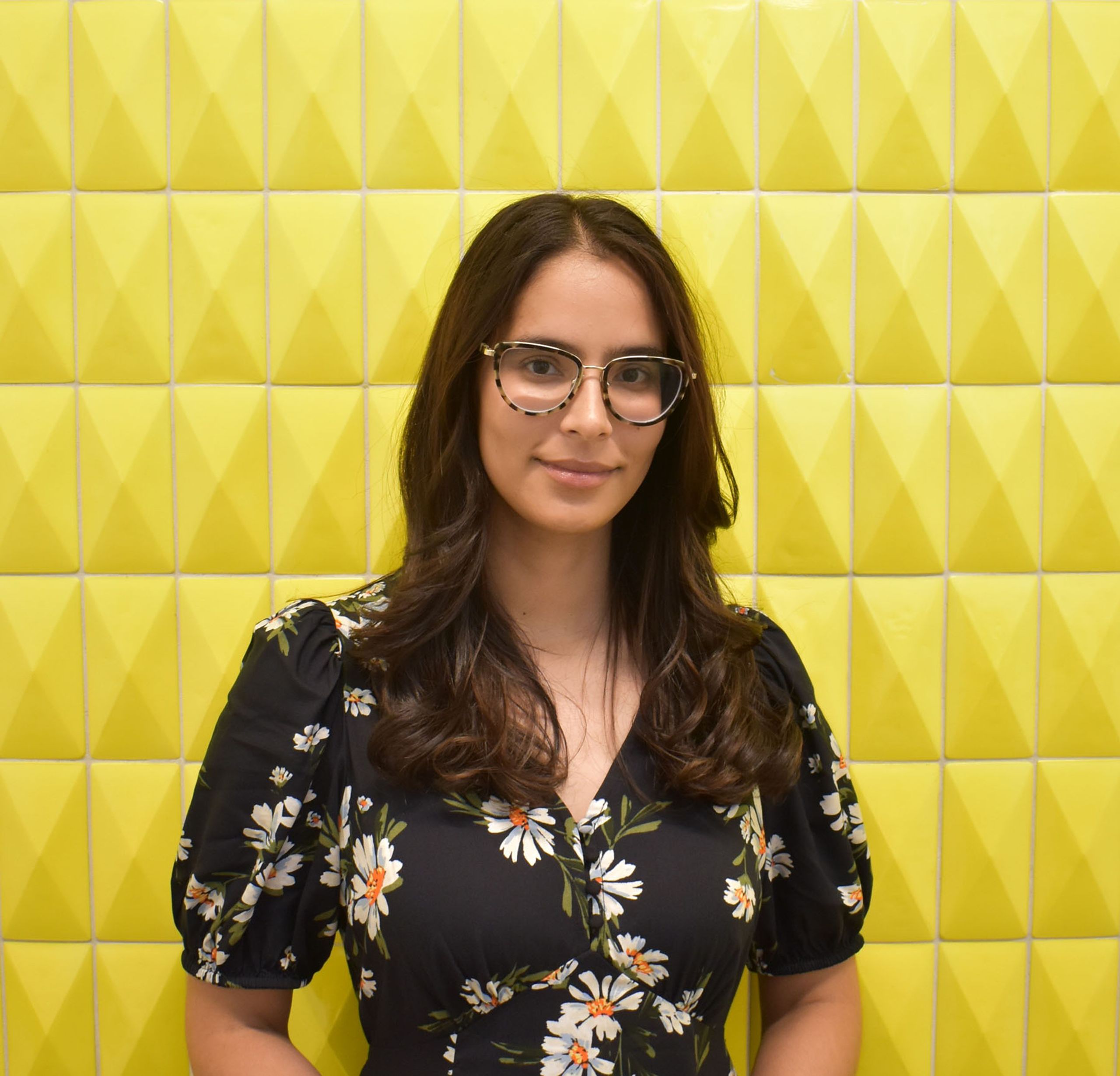 A smiling woman with long dark hair and glasses wears a black floral dress against a textured yellow wall.