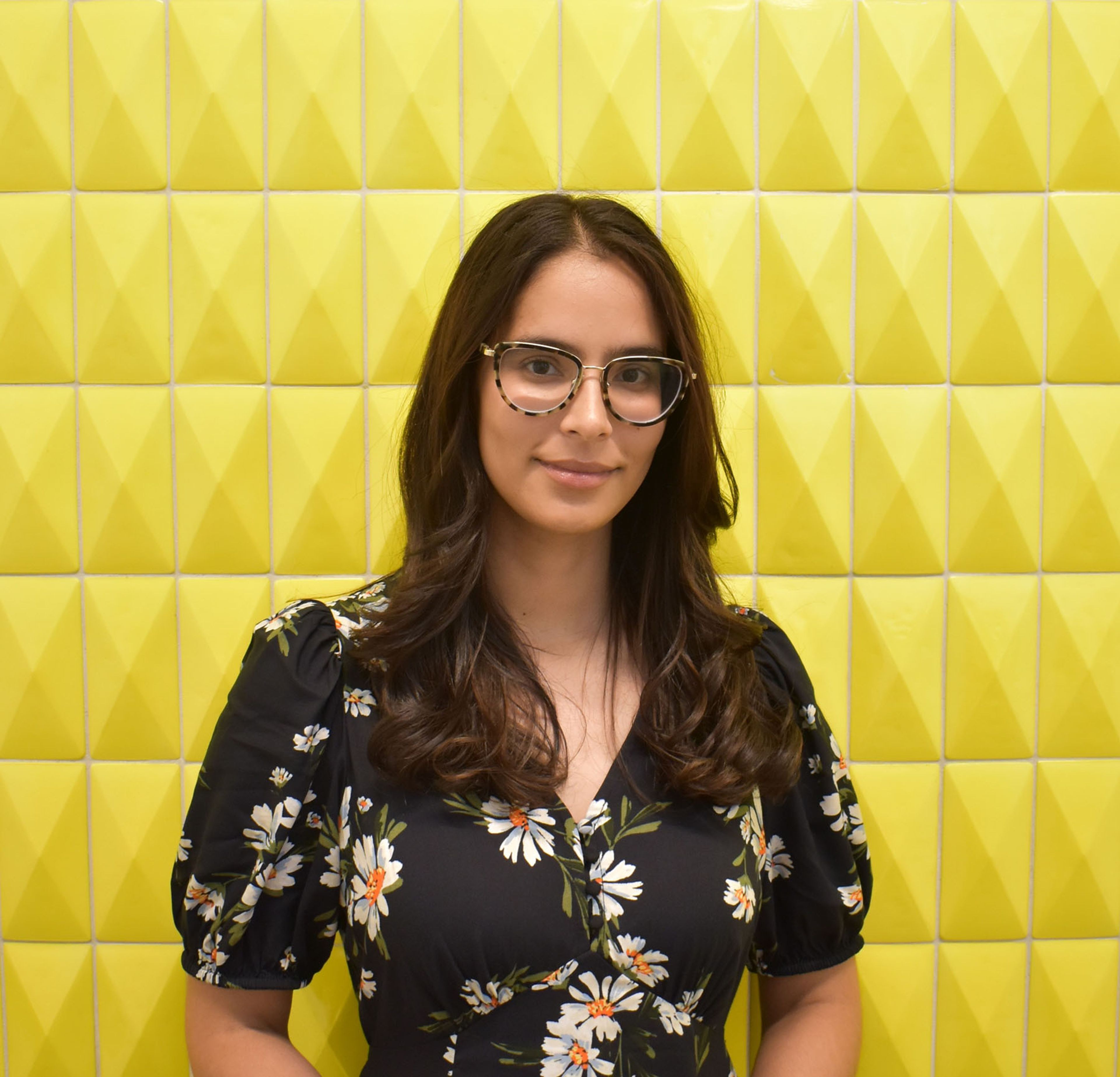 A smiling woman with long dark hair and glasses wears a black floral dress against a textured yellow wall.