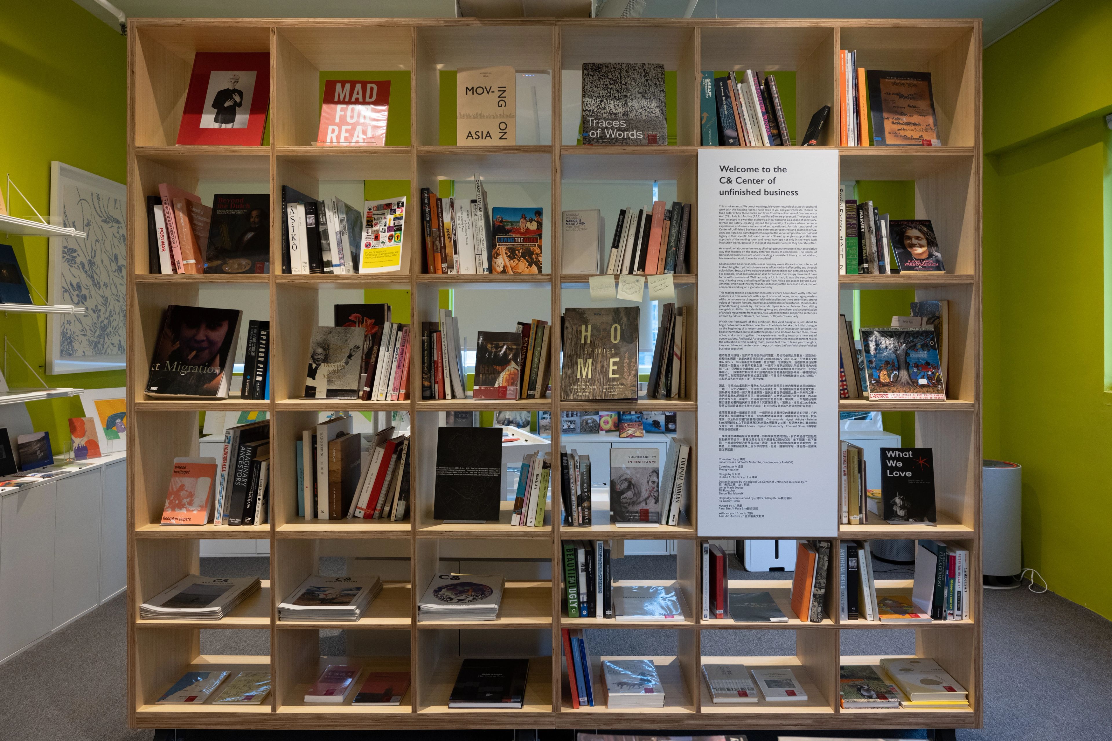 A tall, light wood bookshelf filled with books stands against bright lime green walls, with a banner in the center reading "Welcome to the CA Center of unfinished business".