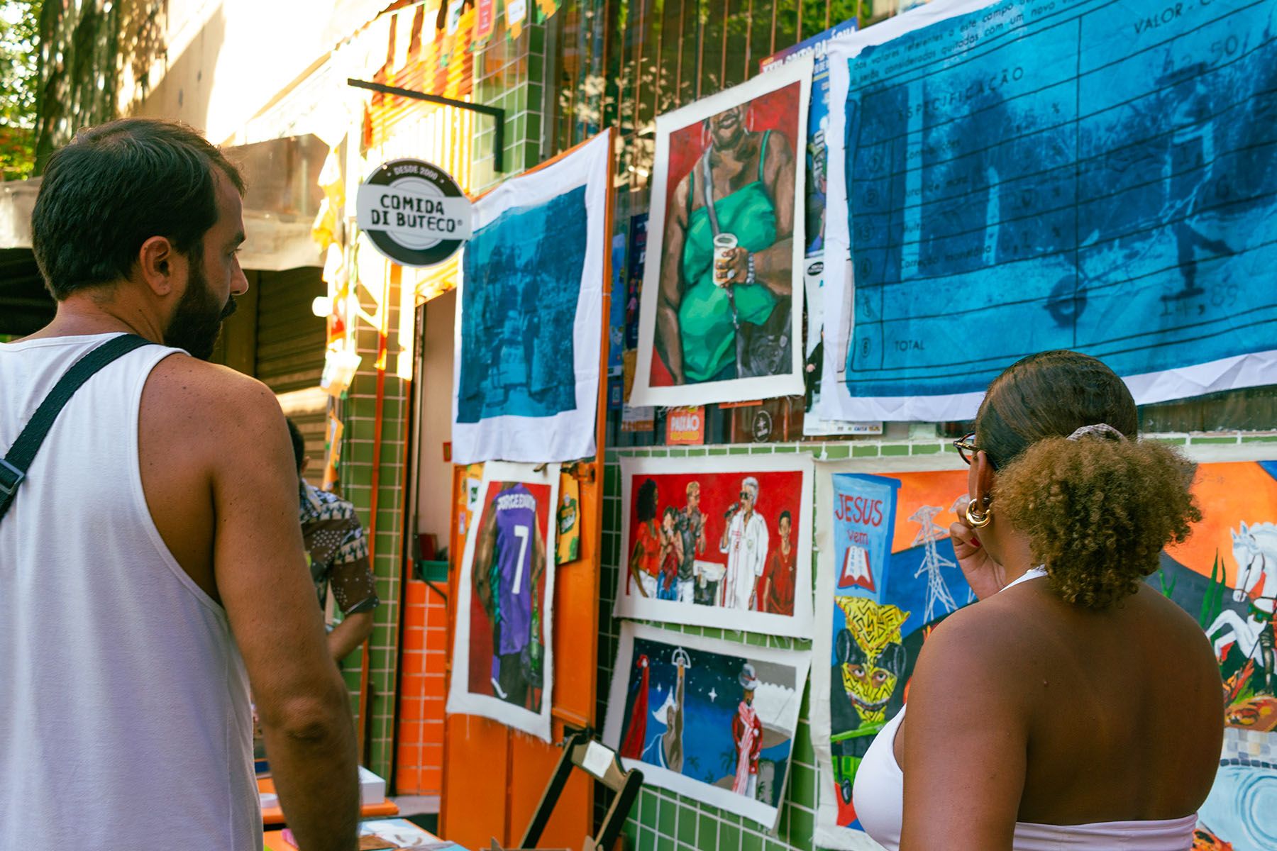 Two people view colorful art and shirts displayed on a green-tiled wall.