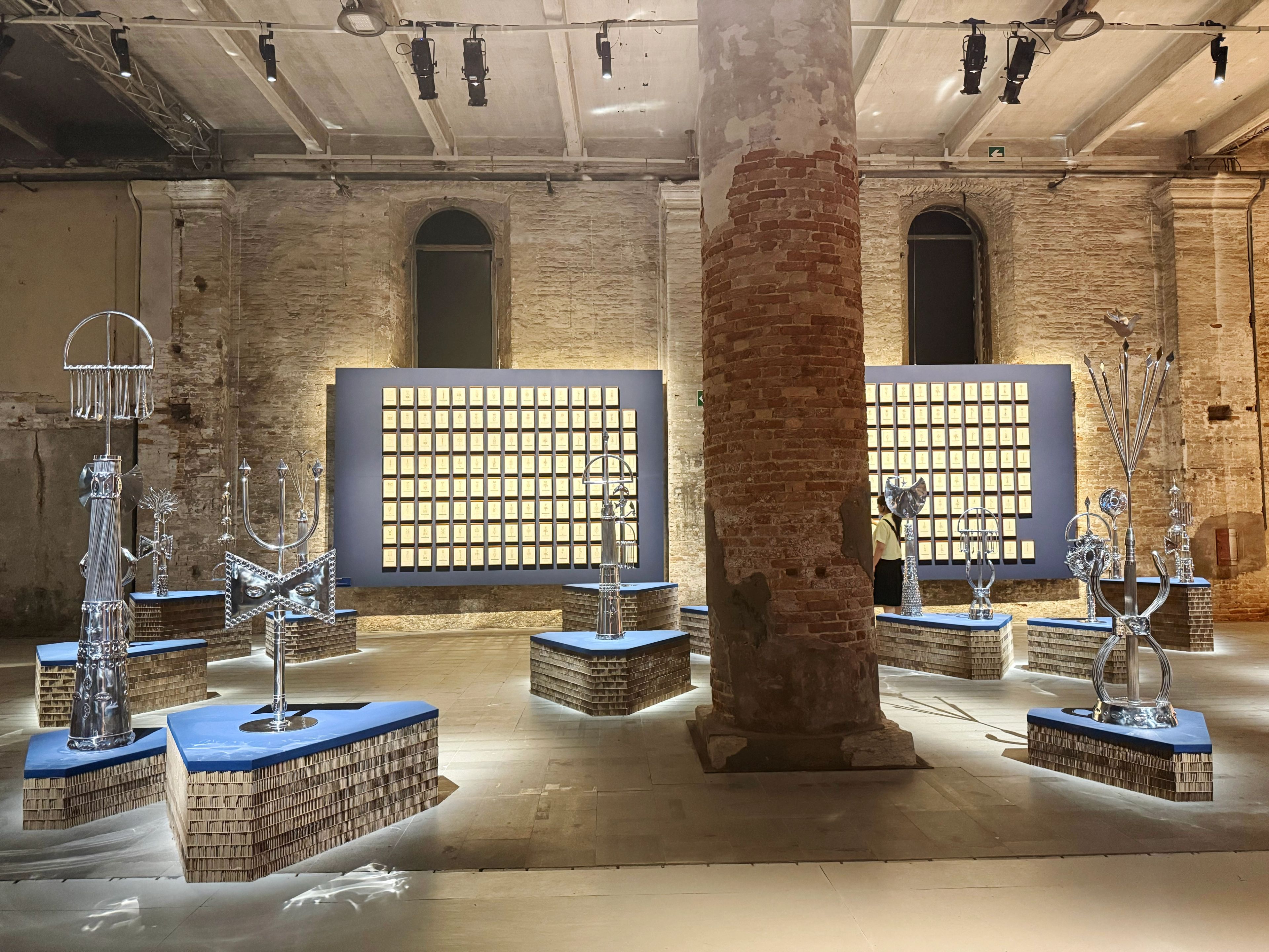An art exhibition in a brick room shows multiple silver abstract sculptures on illuminated geometric bases, and two large dark panels with grids of light-colored squares.