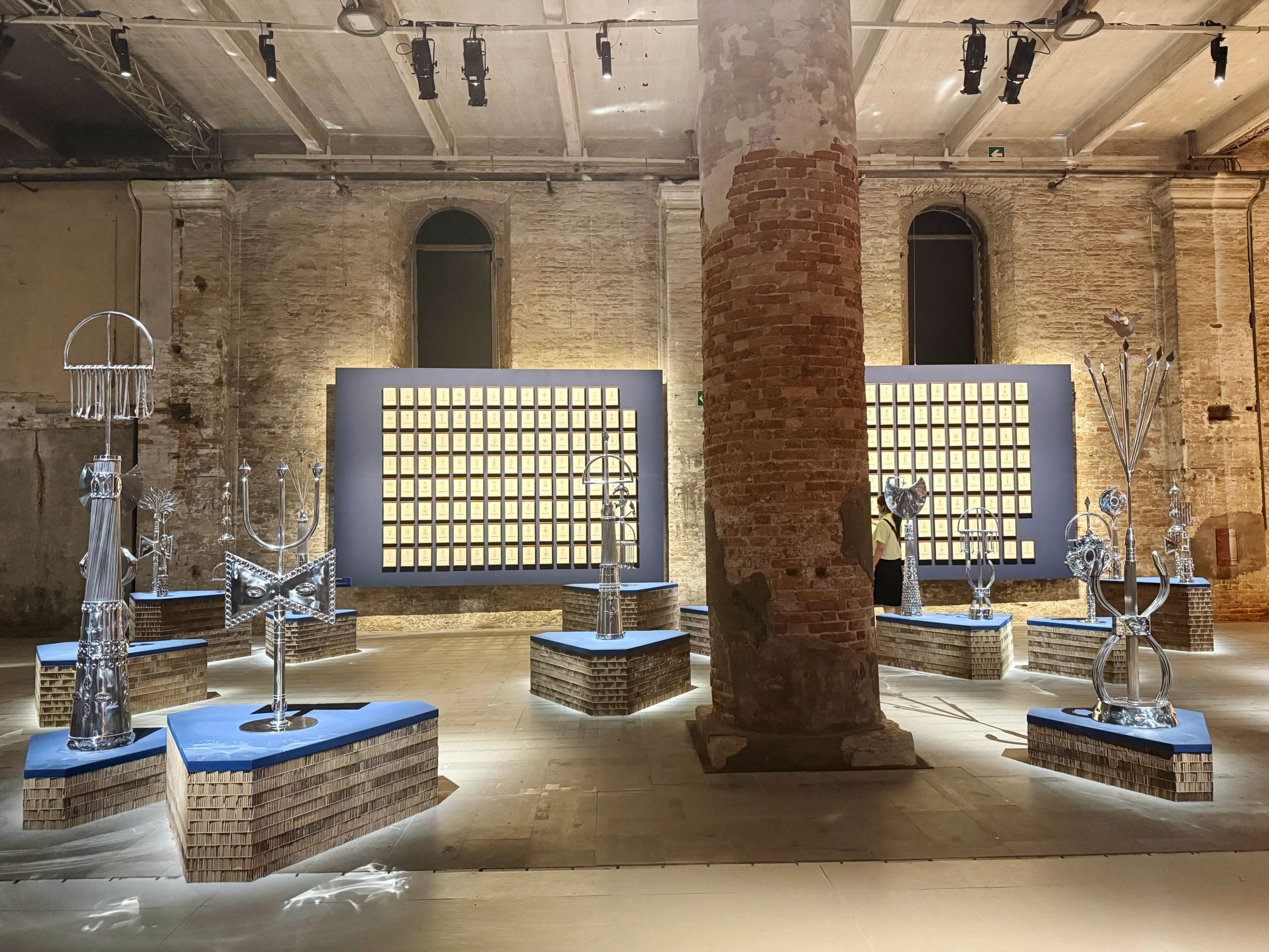 An art exhibition in a brick room shows multiple silver abstract sculptures on illuminated geometric bases, and two large dark panels with grids of light-colored squares.