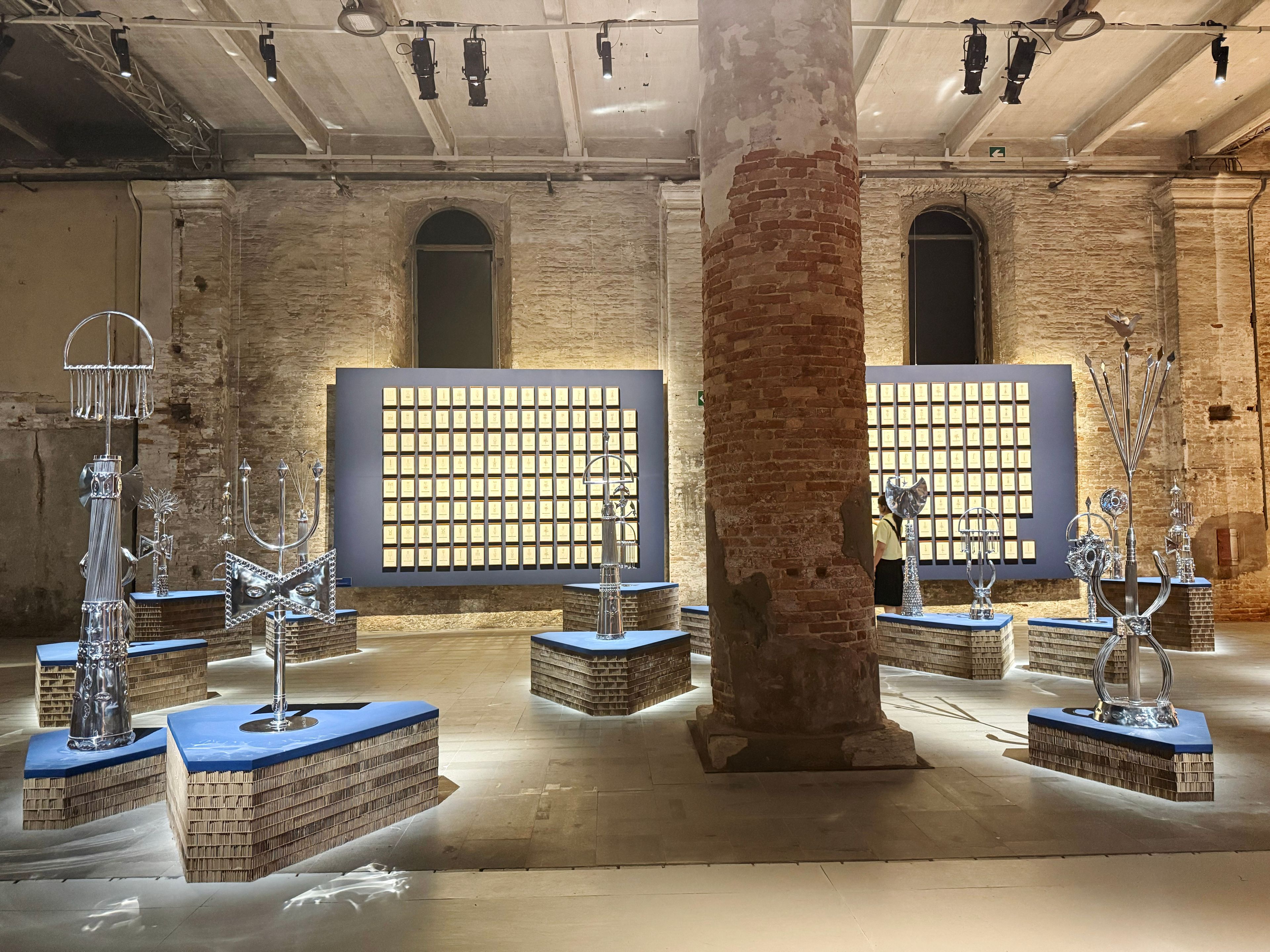 An art exhibition in a brick room shows multiple silver abstract sculptures on illuminated geometric bases, and two large dark panels with grids of light-colored squares.