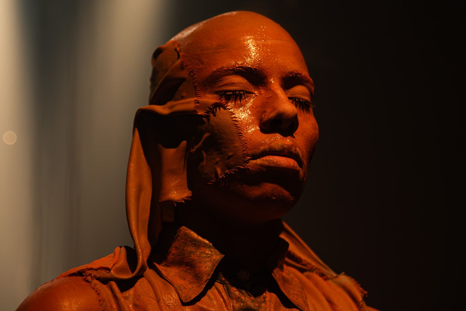 Bald, reddish-brown figure with prominent stitches on the face and closed eyes, wearing a matching collared shirt, dramatically lit.