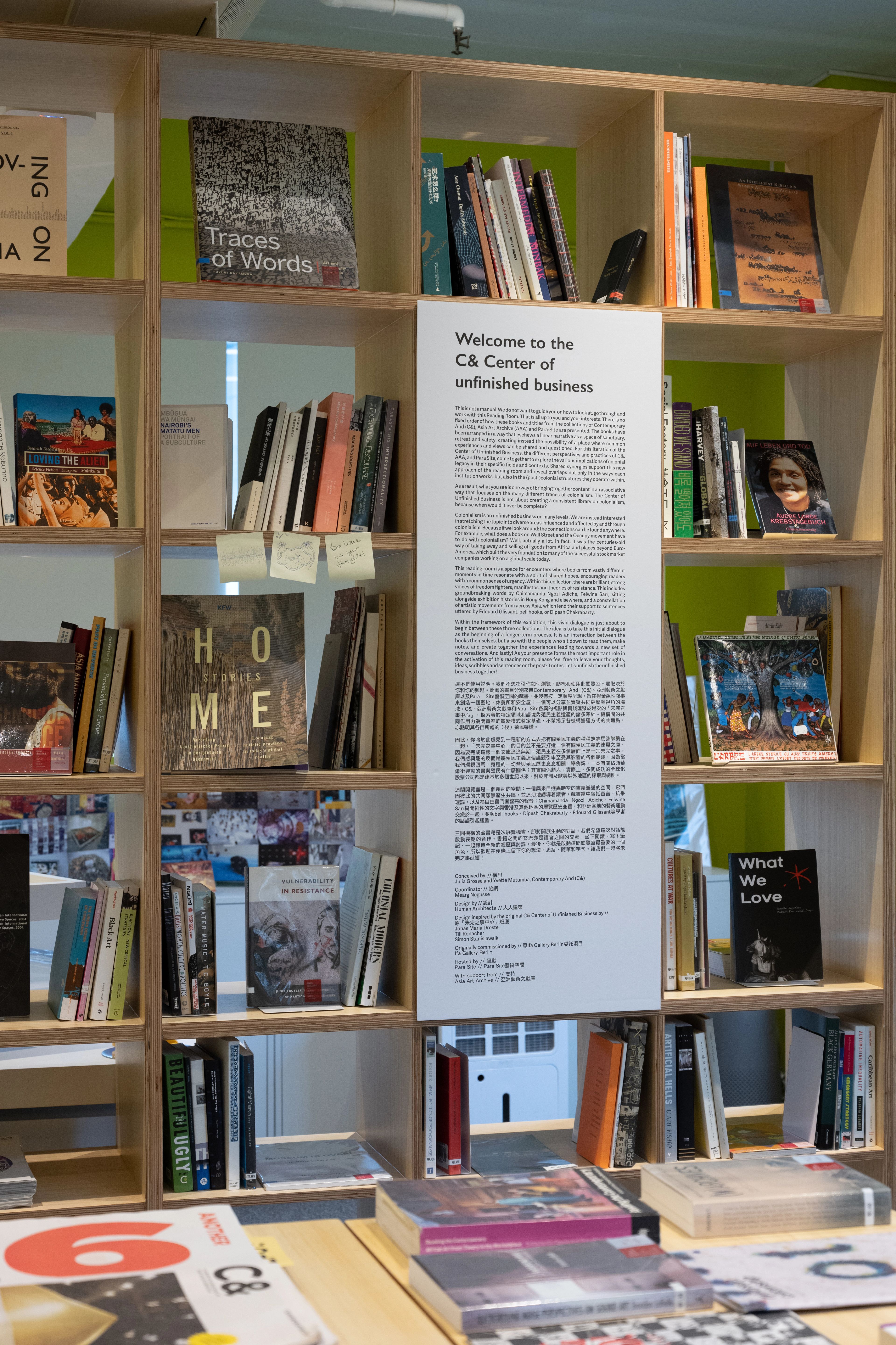 A wooden bookshelf filled with books, displaying a white sign titled "Welcome to the C& Center of unfinished business." Books also lie on a table in the foreground.
