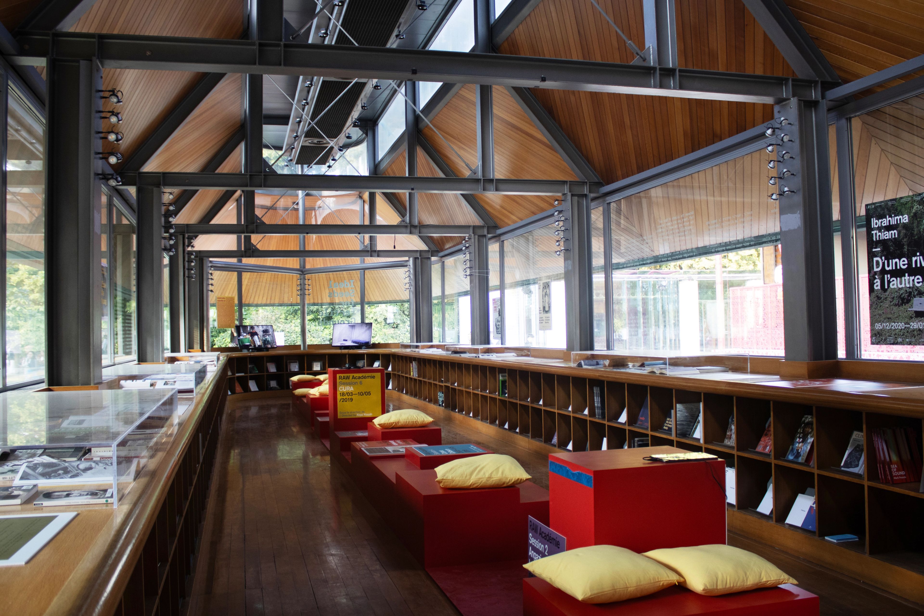 Modern room with a vaulted wood and steel ceiling, large windows, a long shelf of books, and red seating cubes with yellow cushions.