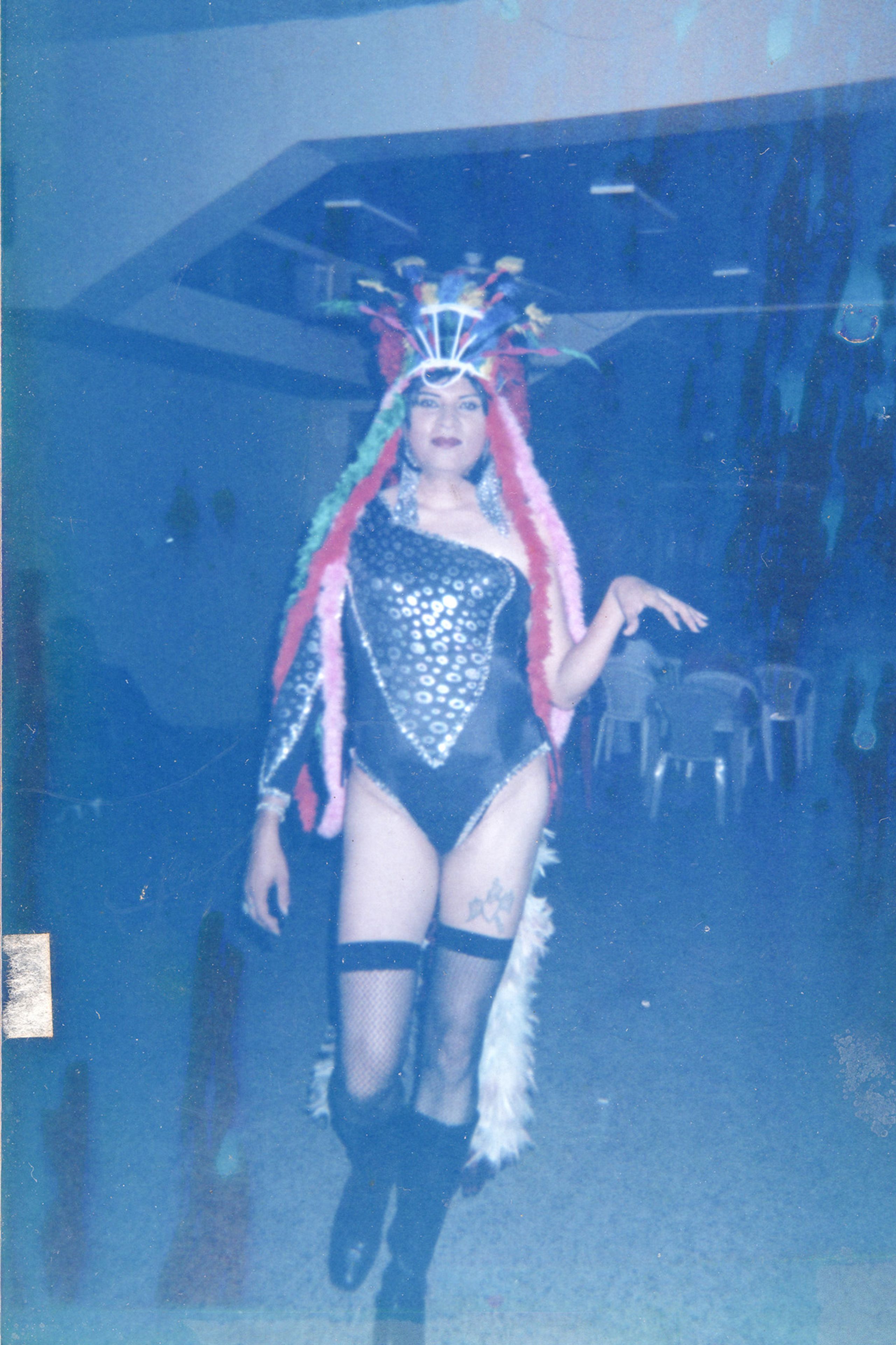 Blue-toned, blurry photograph of a performer in a vibrant drag costume: a large feathered headdress, sequined one-shoulder leotard with white feathery trim, fishnet stockings, and boots.