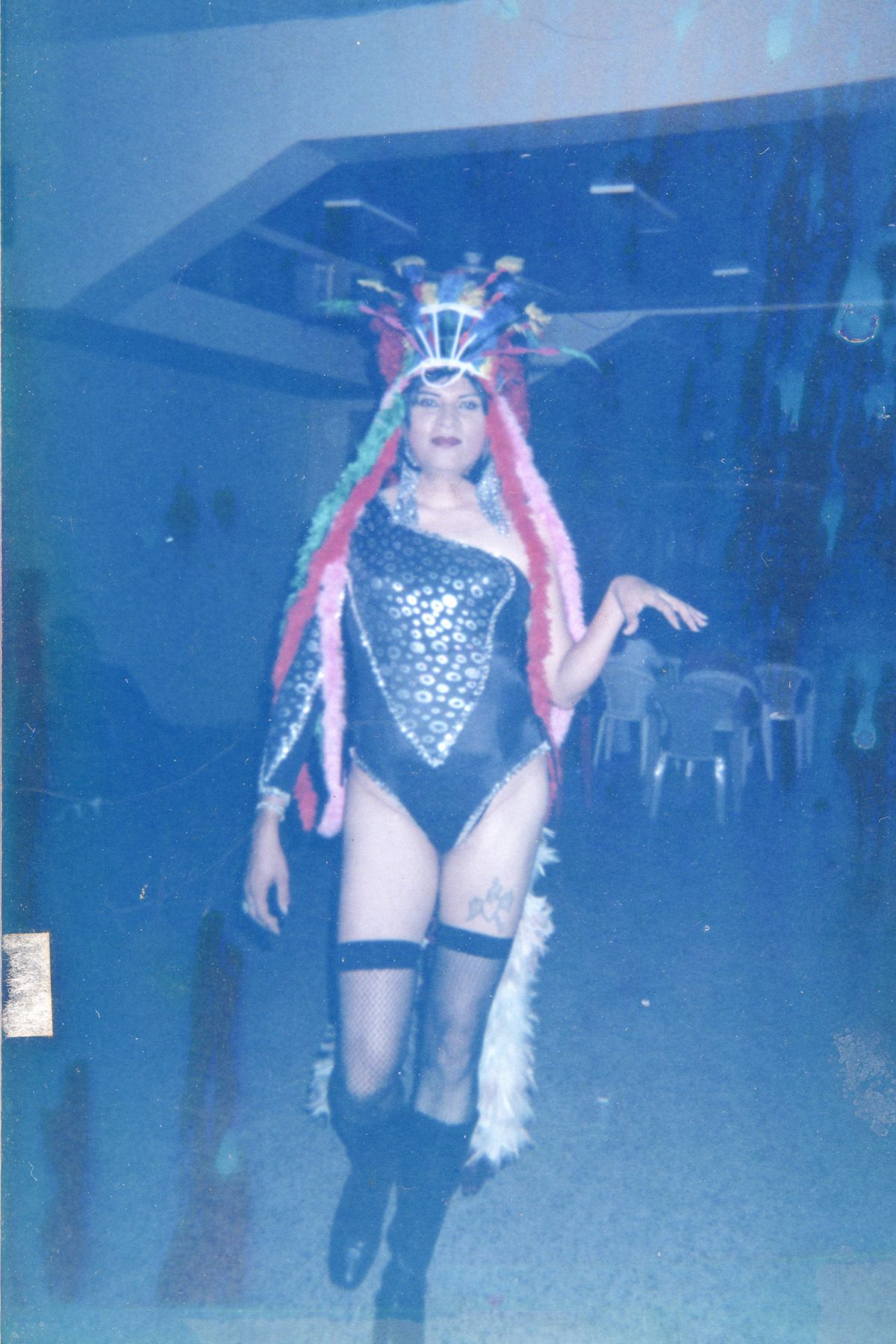 Blue-toned, blurry photograph of a performer in a vibrant drag costume: a large feathered headdress, sequined one-shoulder leotard with white feathery trim, fishnet stockings, and boots.