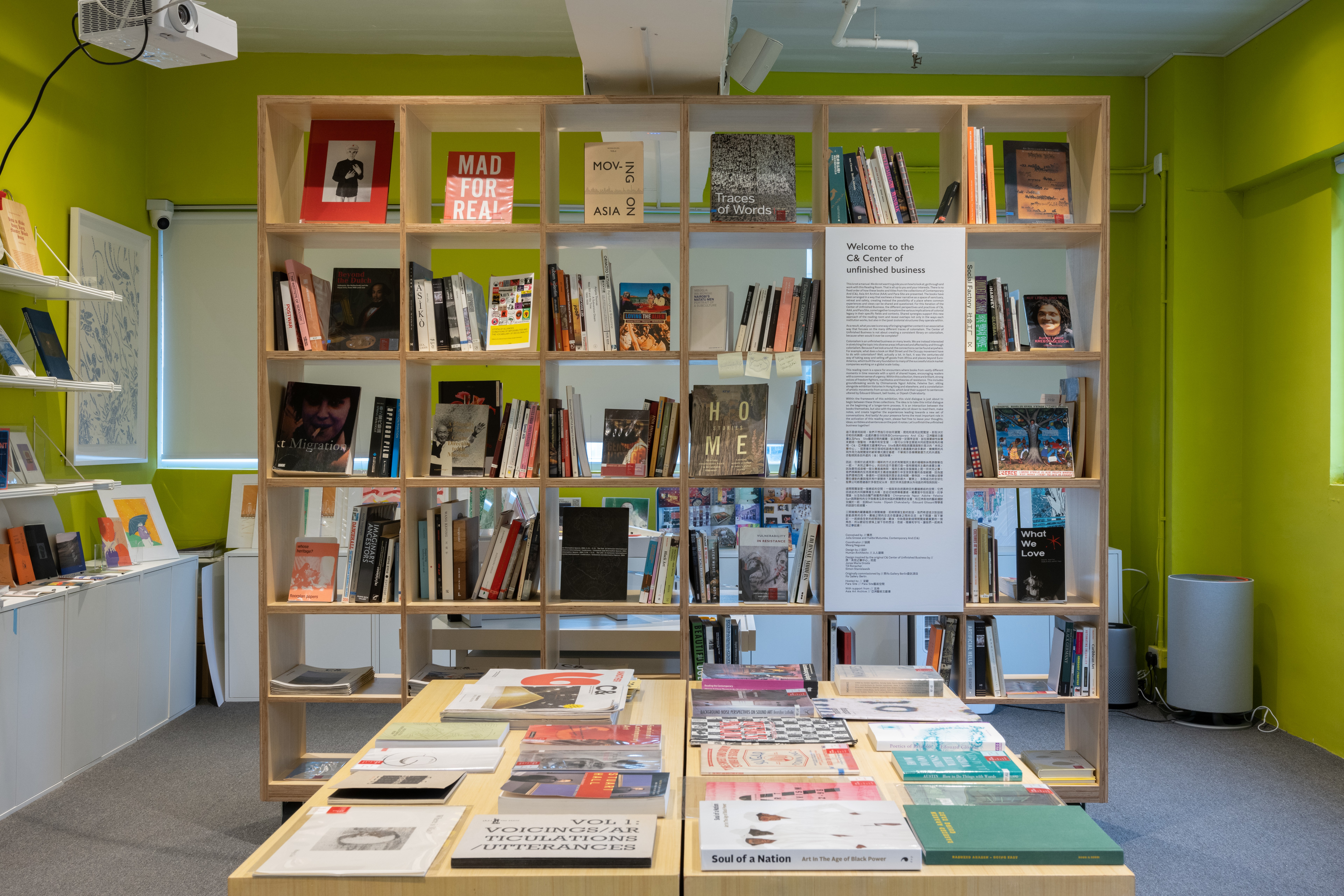 A lime green room with a large wooden bookshelf packed with books, a table displaying more books, and a white informational panel.