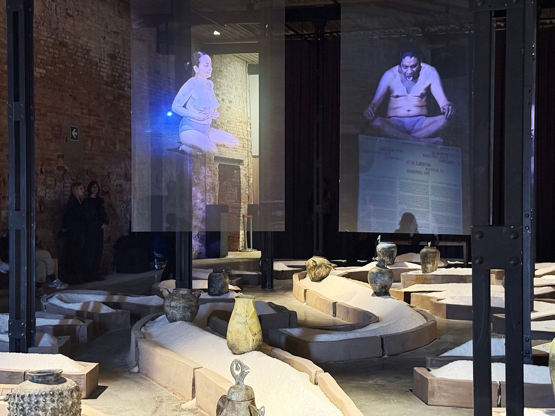 An art exhibition features two large projections of people meditating, above winding sand beds containing various ceramic vessels.